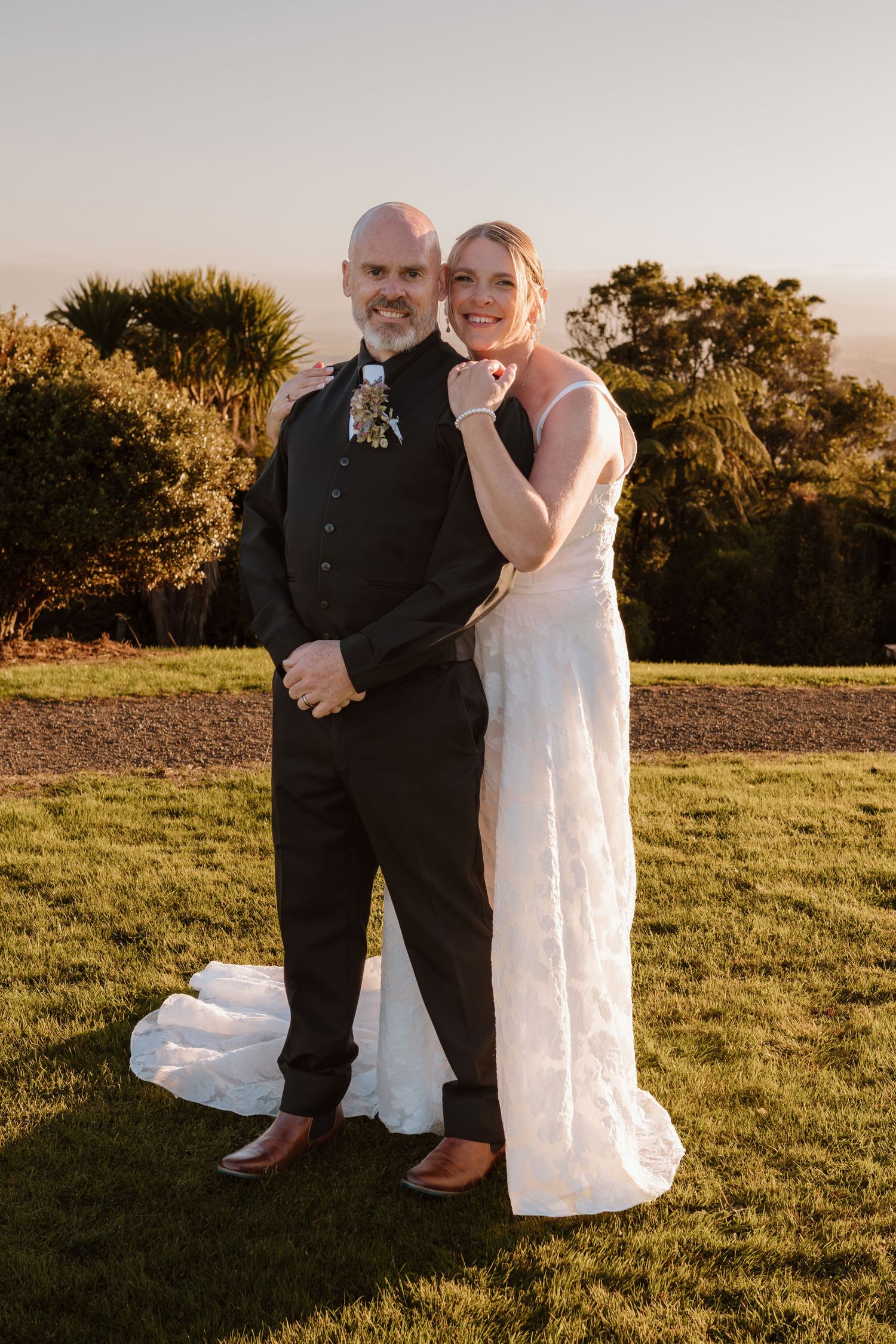 A couple in wedding attire sits together on grassy field during golden hour sunset.