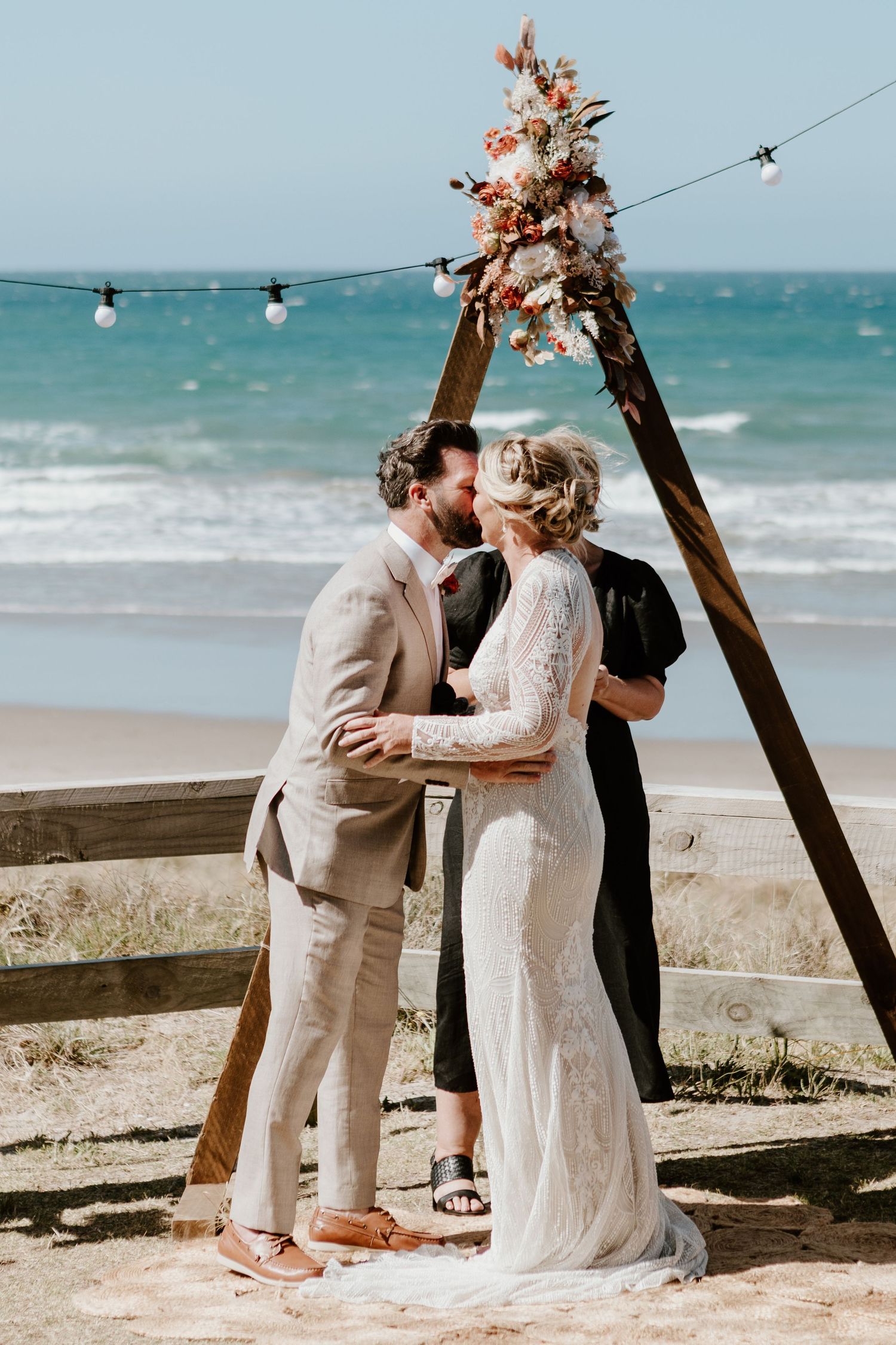 A beachside wedding ceremony under a wooden arch decorated with pink flowers and string lights by the ocean.