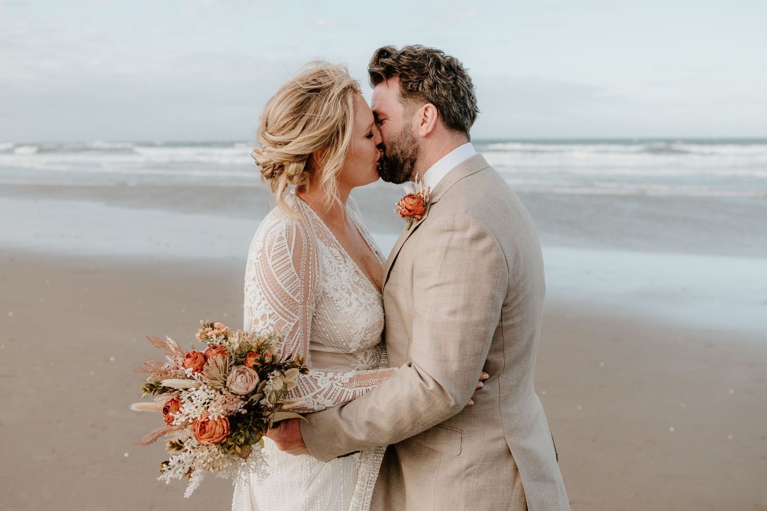 Romantic beach wedding couple embrace while holding coral-colored bouquet on misty oceanfront.