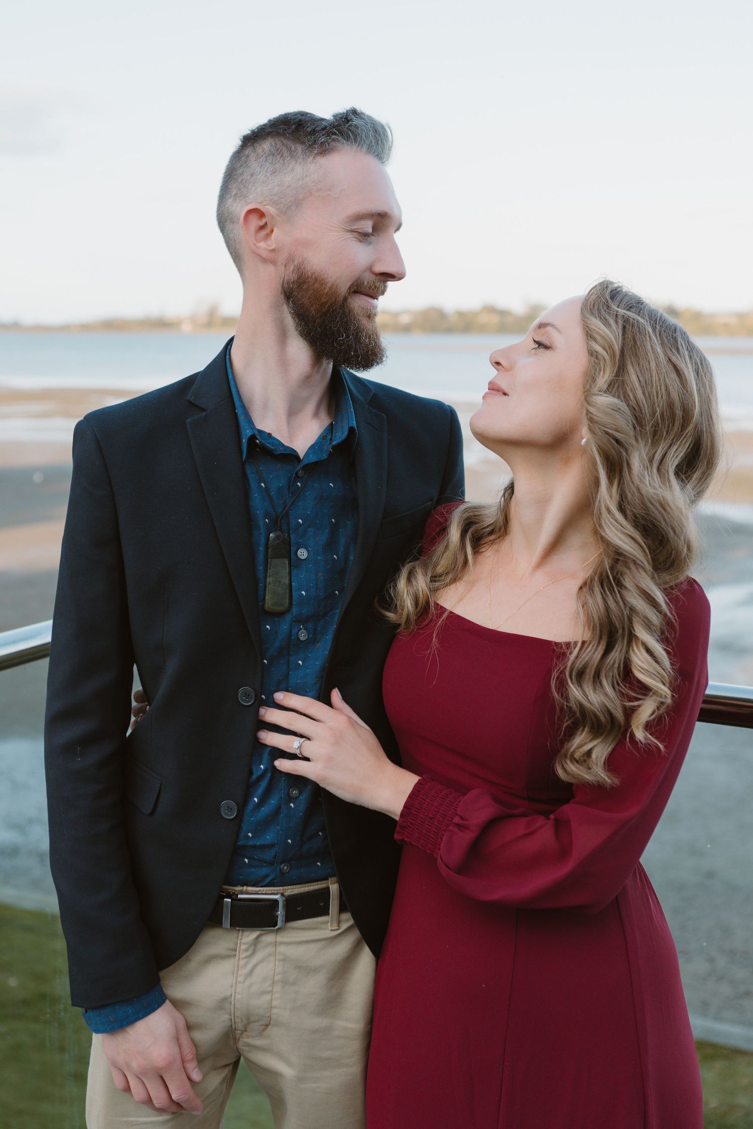 A romantic couple in formal attire poses together at sunset, with the woman in burgundy and man in navy and khaki.