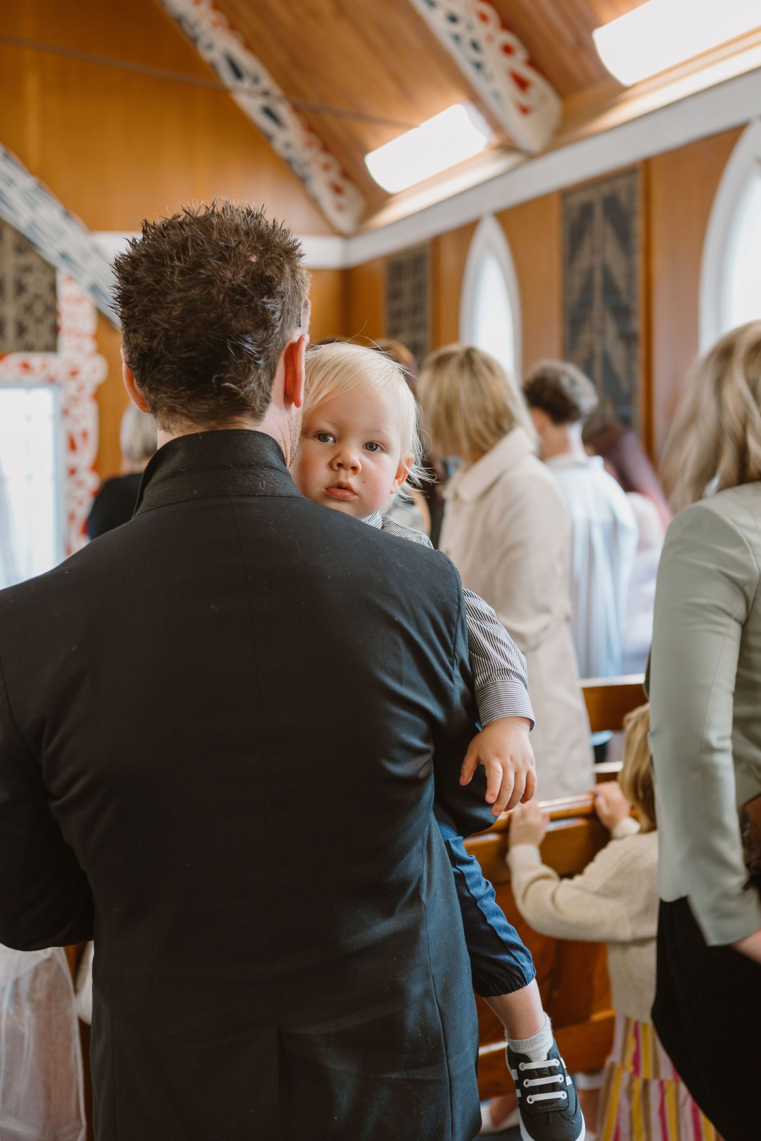 A person in a dark suit facing away from the camera holds a baby in a casual indoor setting.