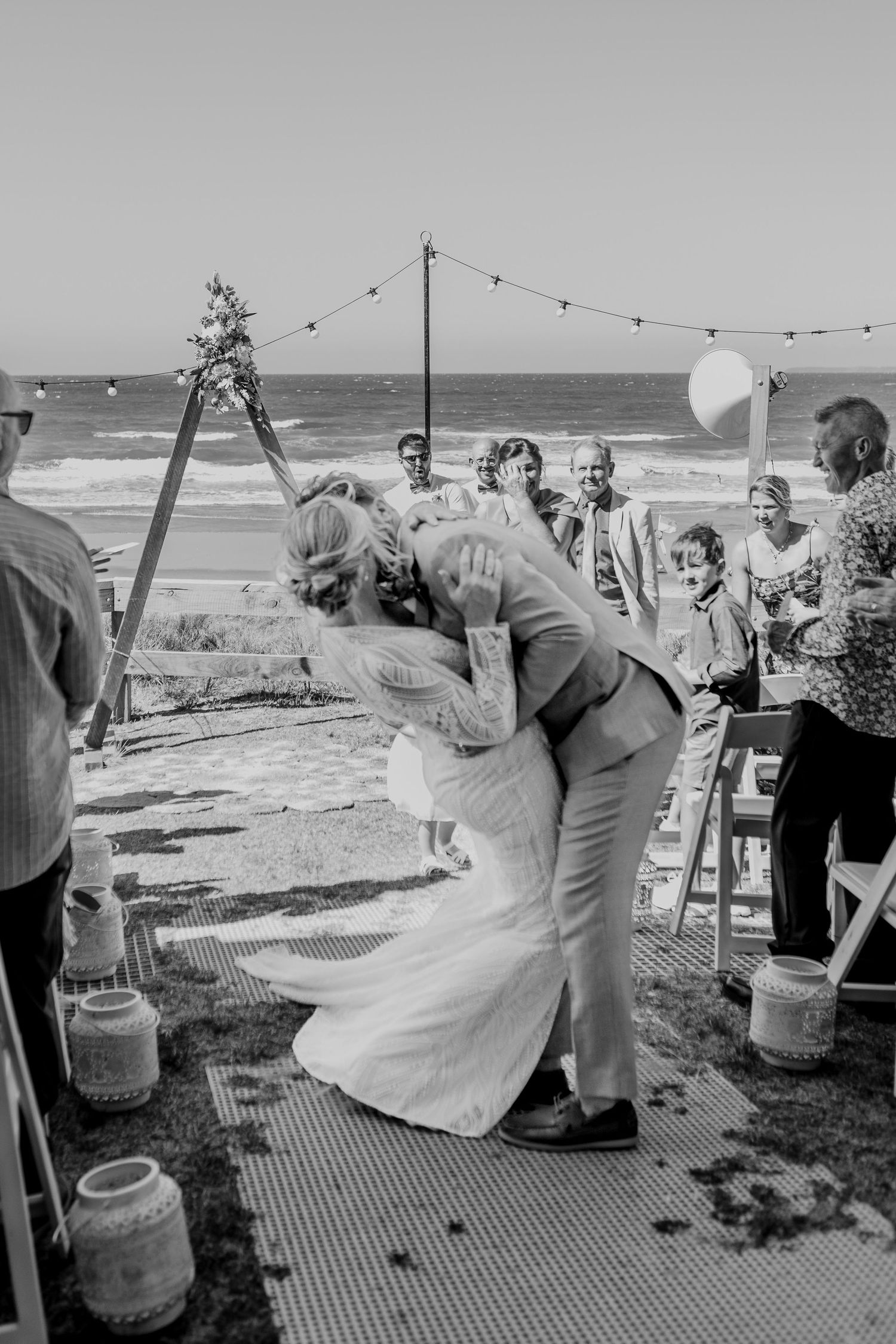 Black and white photo of wedding guests dancing at outdoor beach reception. Dip kiss Papamoa Beach Wedding