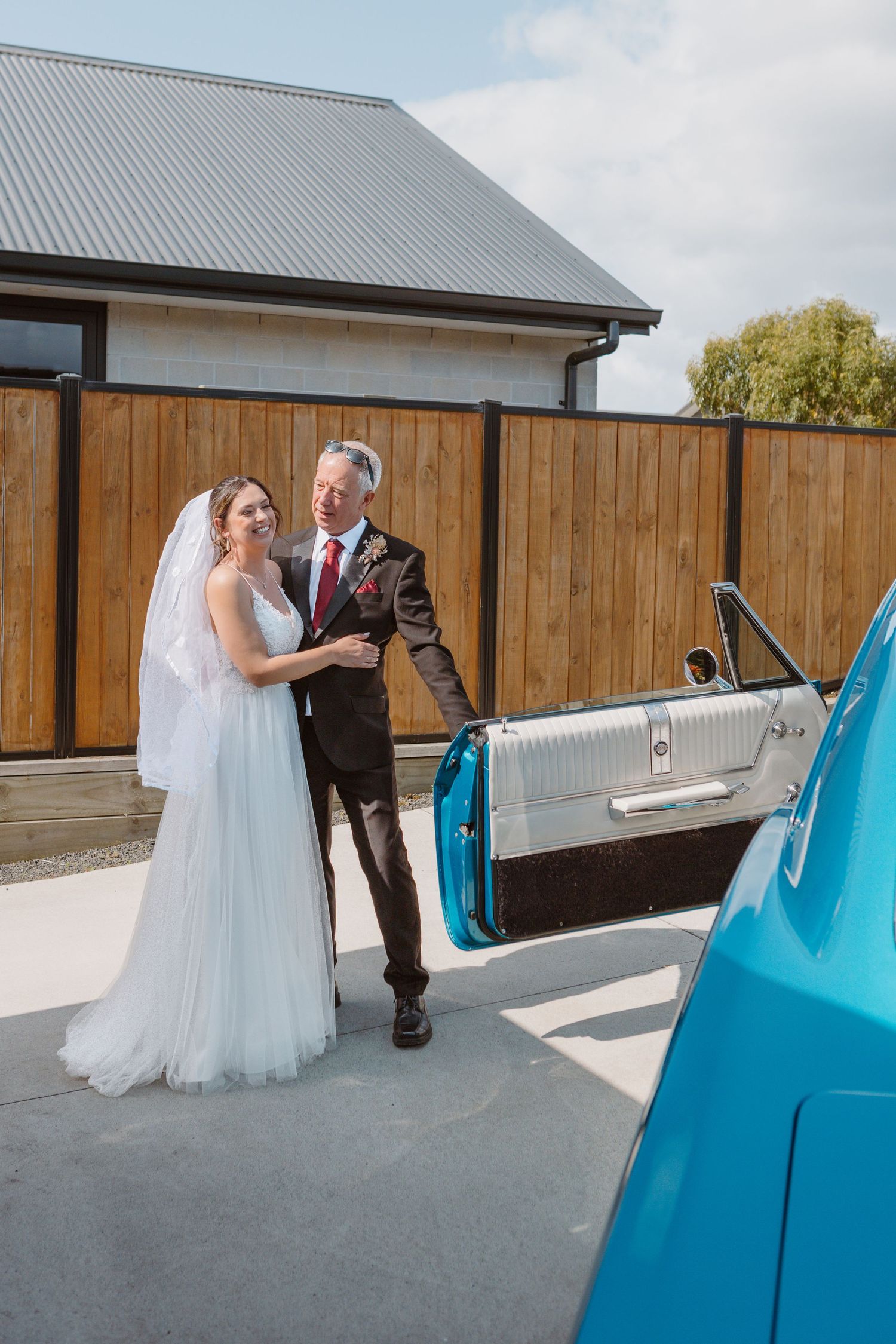 A couple poses by a vintage turquoise car on their wedding day with wooden fence and building in background.