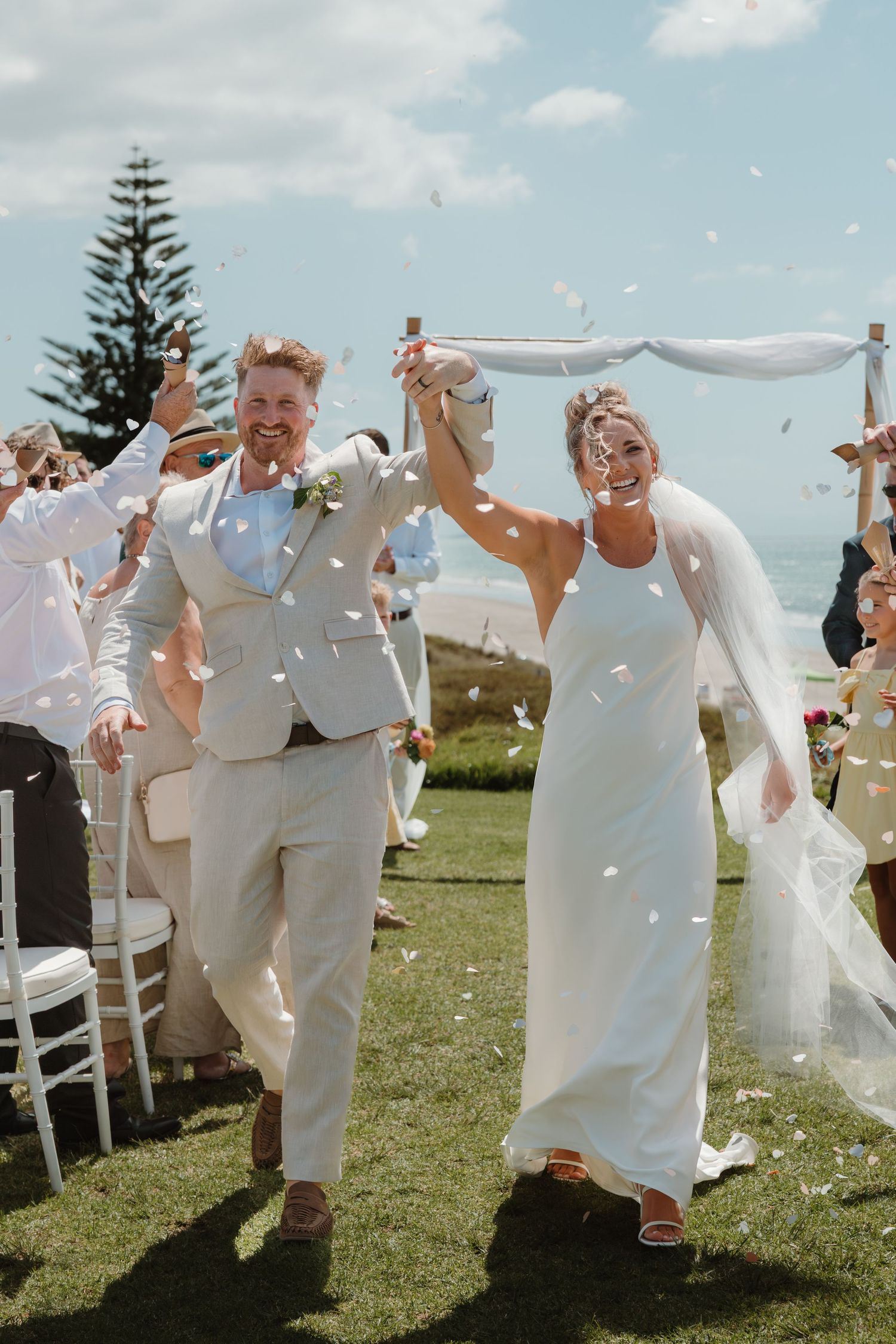 Joyful newlyweds walk down the aisle as confetti fills the air at an outdoor seaside wedding ceremony.