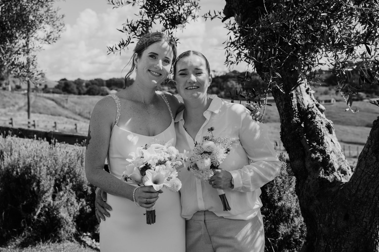 Two brides pose together outdoors holding white bouquets on their wedding day in a black and white photo.