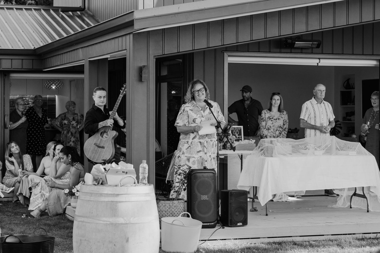 Woman speaks at outdoor event with band and guests at a rustic venue in black and white photo.