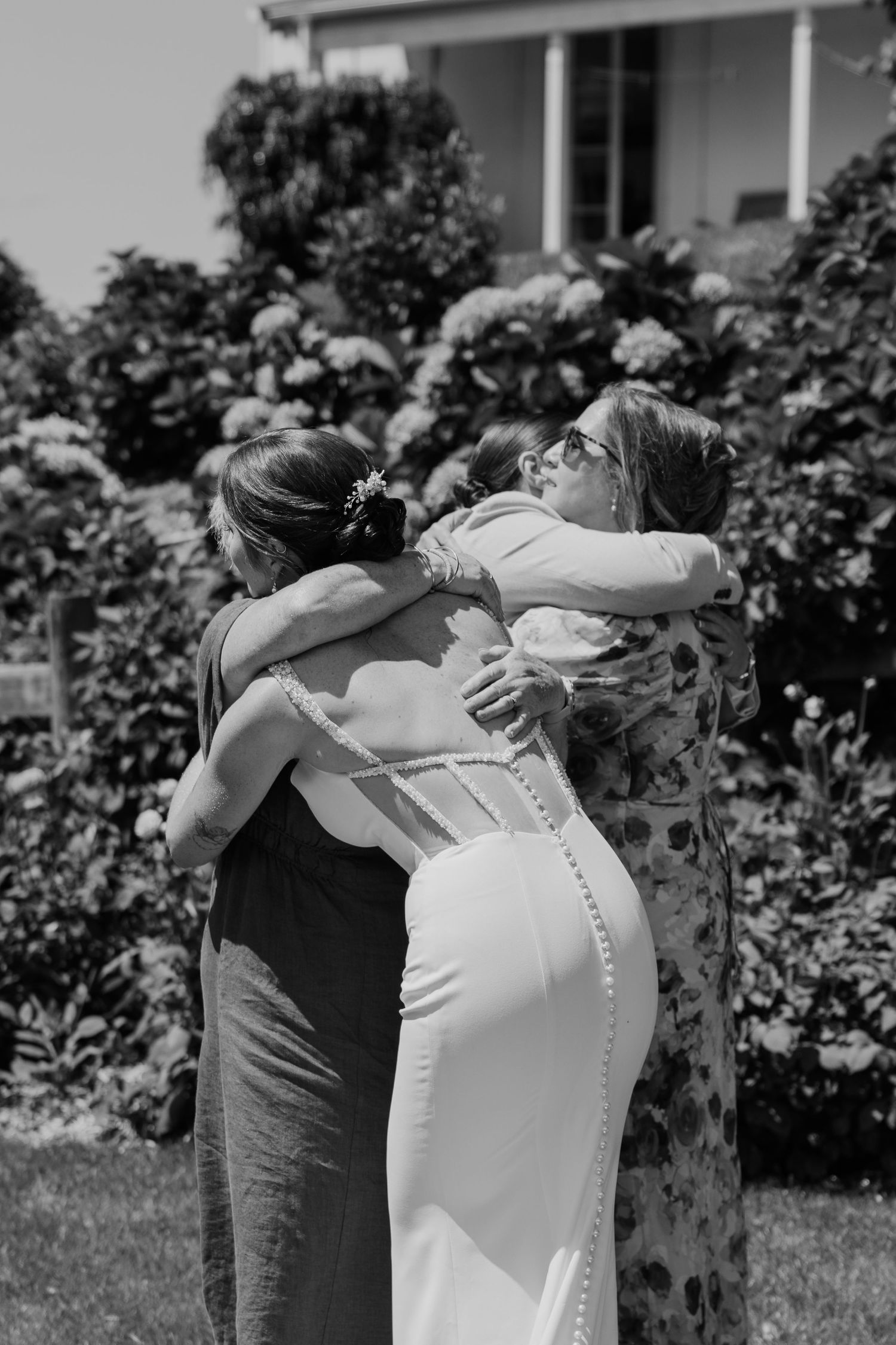 Black and white photo of a bride in white gown sharing a group hug with two women in a garden setting.