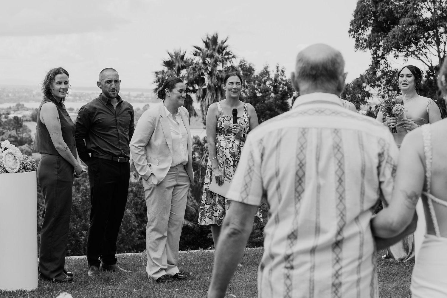 Black and white outdoor wedding gathering with guests socializing on a hilltop with palm trees and city views.