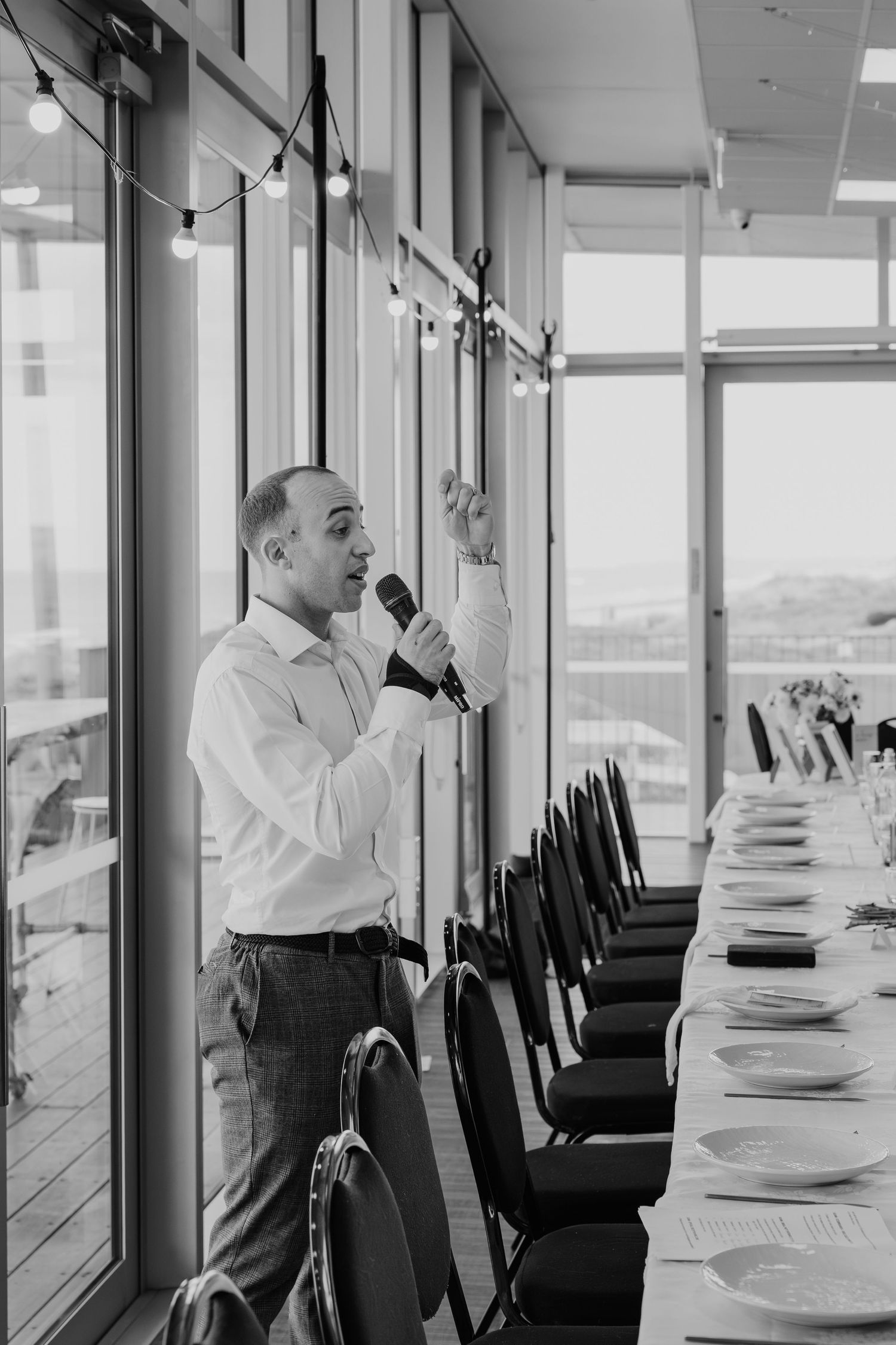 Man in white shirt speaking into microphone at elegant banquet hall with long dining table and large windows.