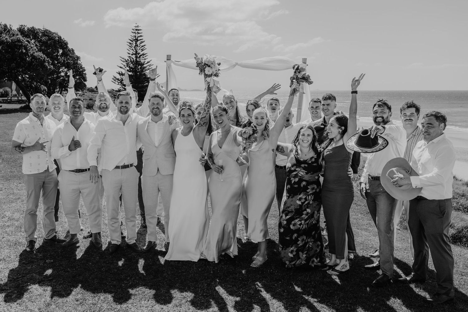 Black and white wedding party photo with bride, groom, and guests celebrating outdoors near the ocean.