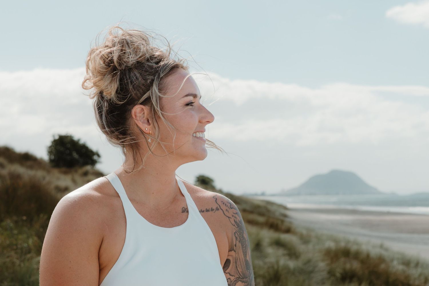 Woman with tattoo and hair bun smiling at beach landscape on cloudy day.