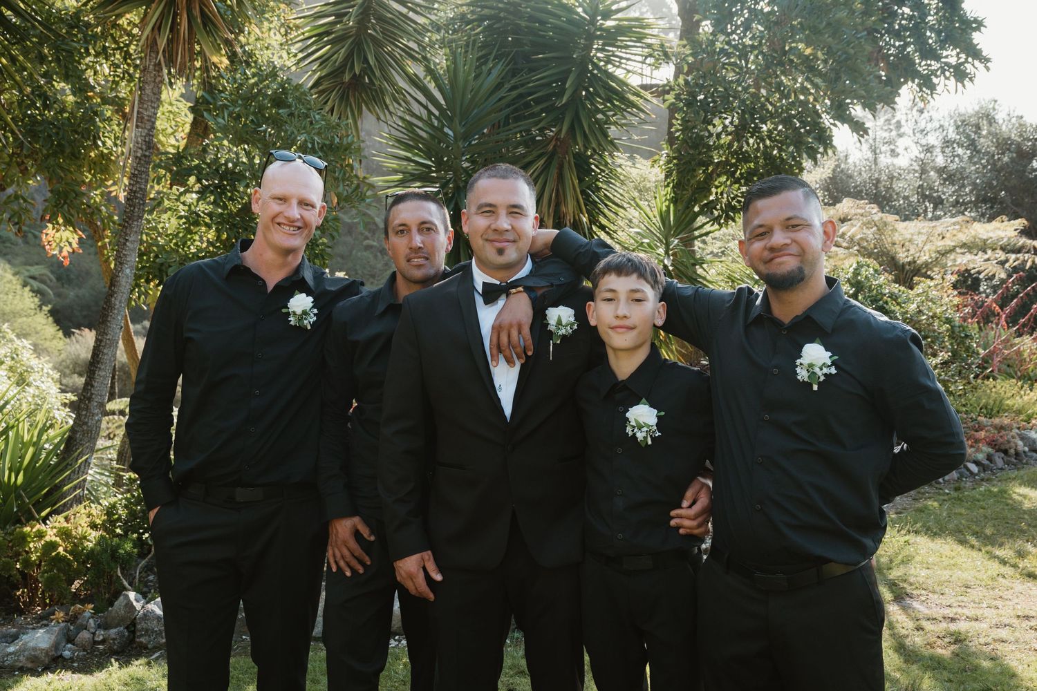 Wedding party in black suits poses together outdoors in a garden setting with palm trees and greenery.