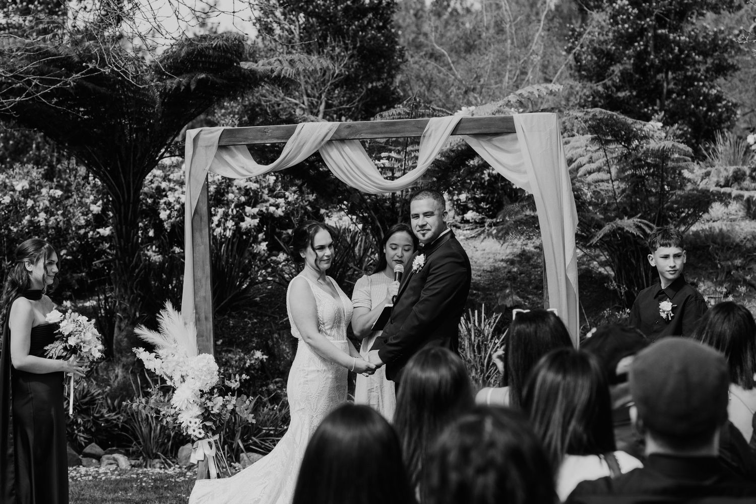 Wedding ceremony takes place under a white canopy in an outdoor garden setting with guests seated on white chairs.