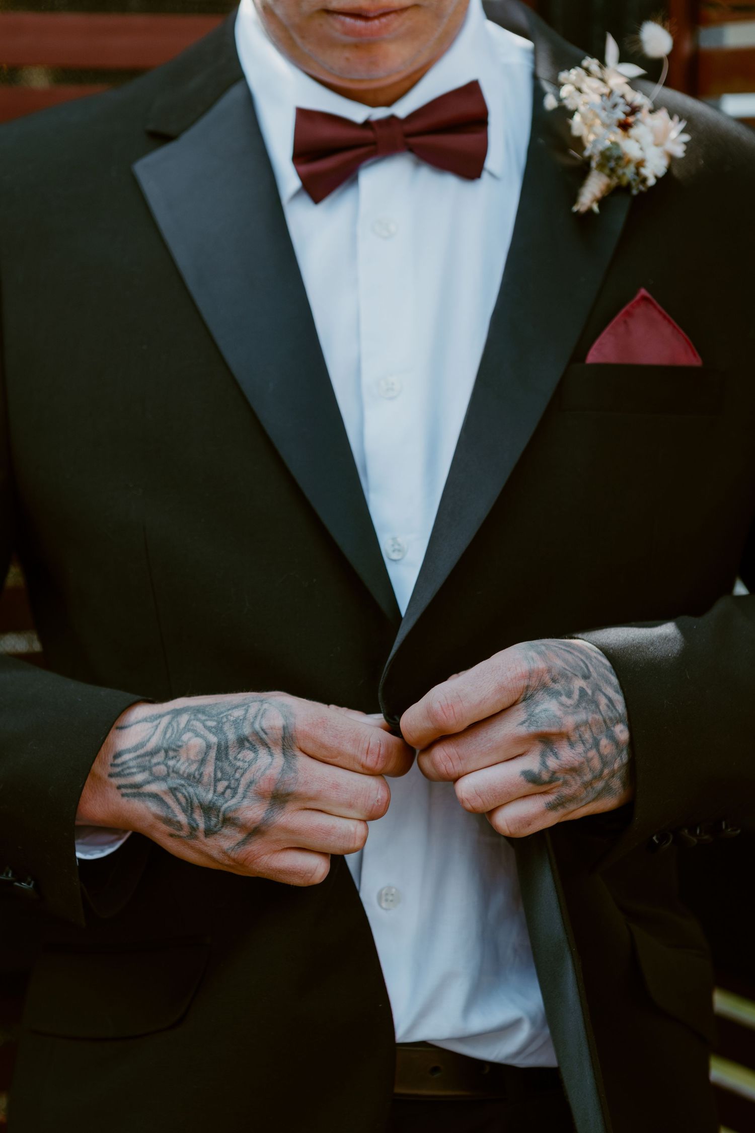 Closeup of groom in black tuxedo with burgundy bow tie and boutonniere showing tattooed hands buttoning jacket.