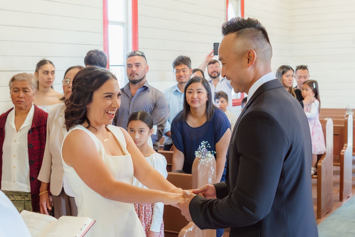 A joyful couple exchanges rings during their wedding ceremony in a bright church sanctuary as guests look on.
