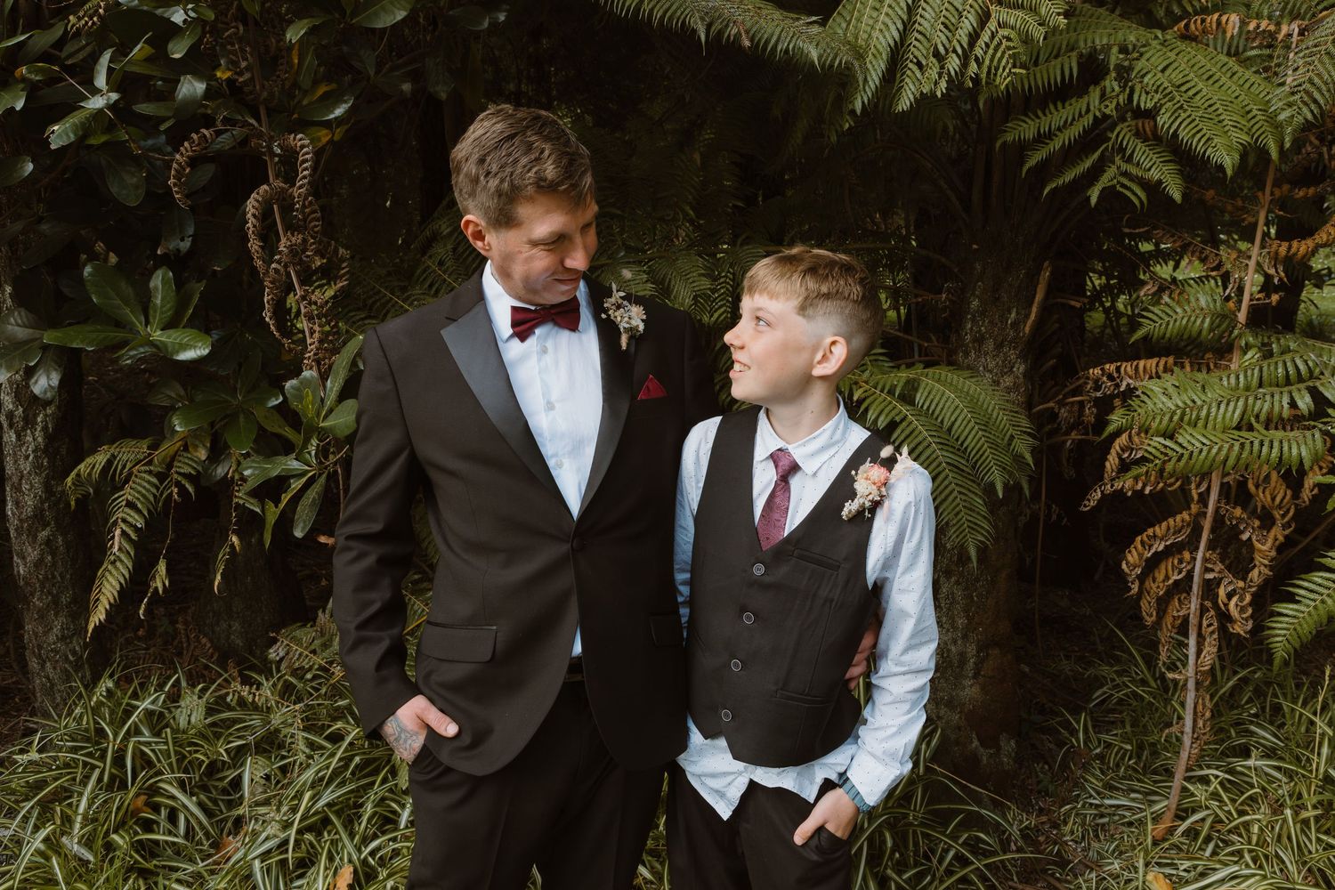 A father and son share a tender candid moment together amid lush greenery in formal attire during a wedding celebration.