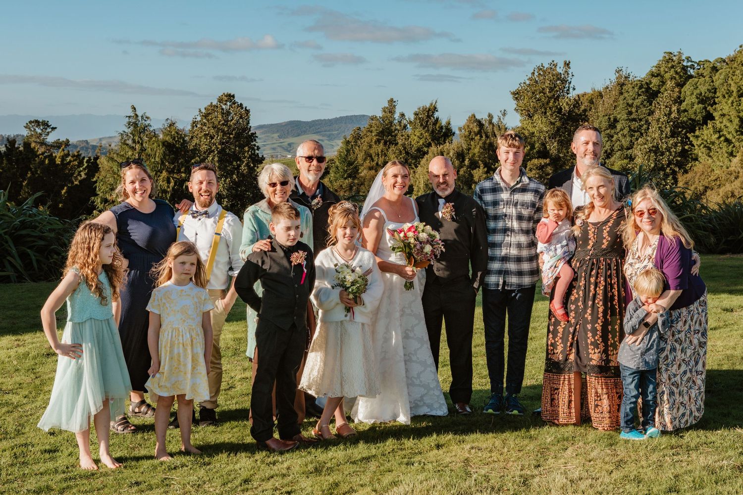 Group of people gathered outdoors for a wedding celebration on a grassy hillside with trees and mountains in the background.