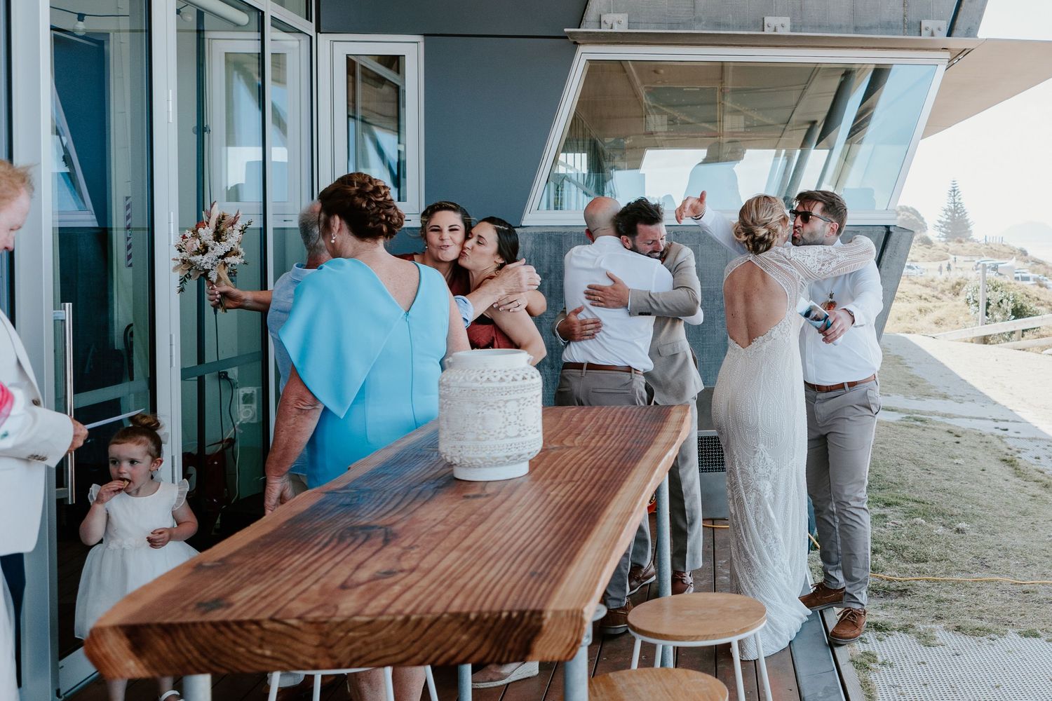 Wedding guests gather around a wooden table on an outdoor deck. Papamoa Surf Lifesaving Club Wedding