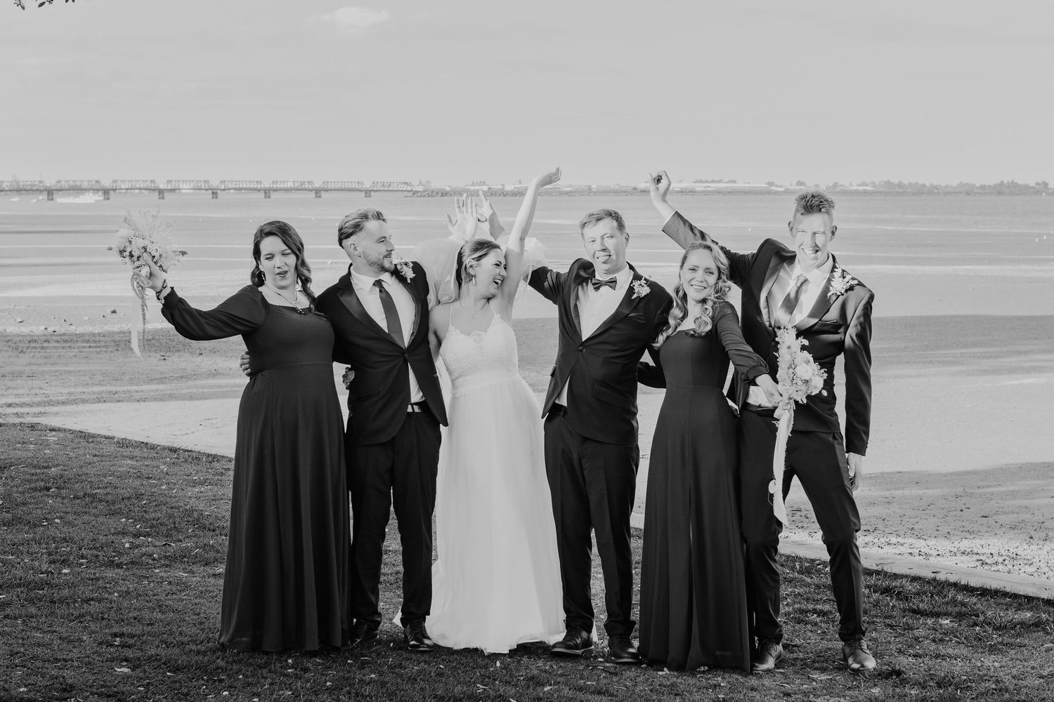 Wedding party celebrates with raised arms in a joyful group photo by the water.
