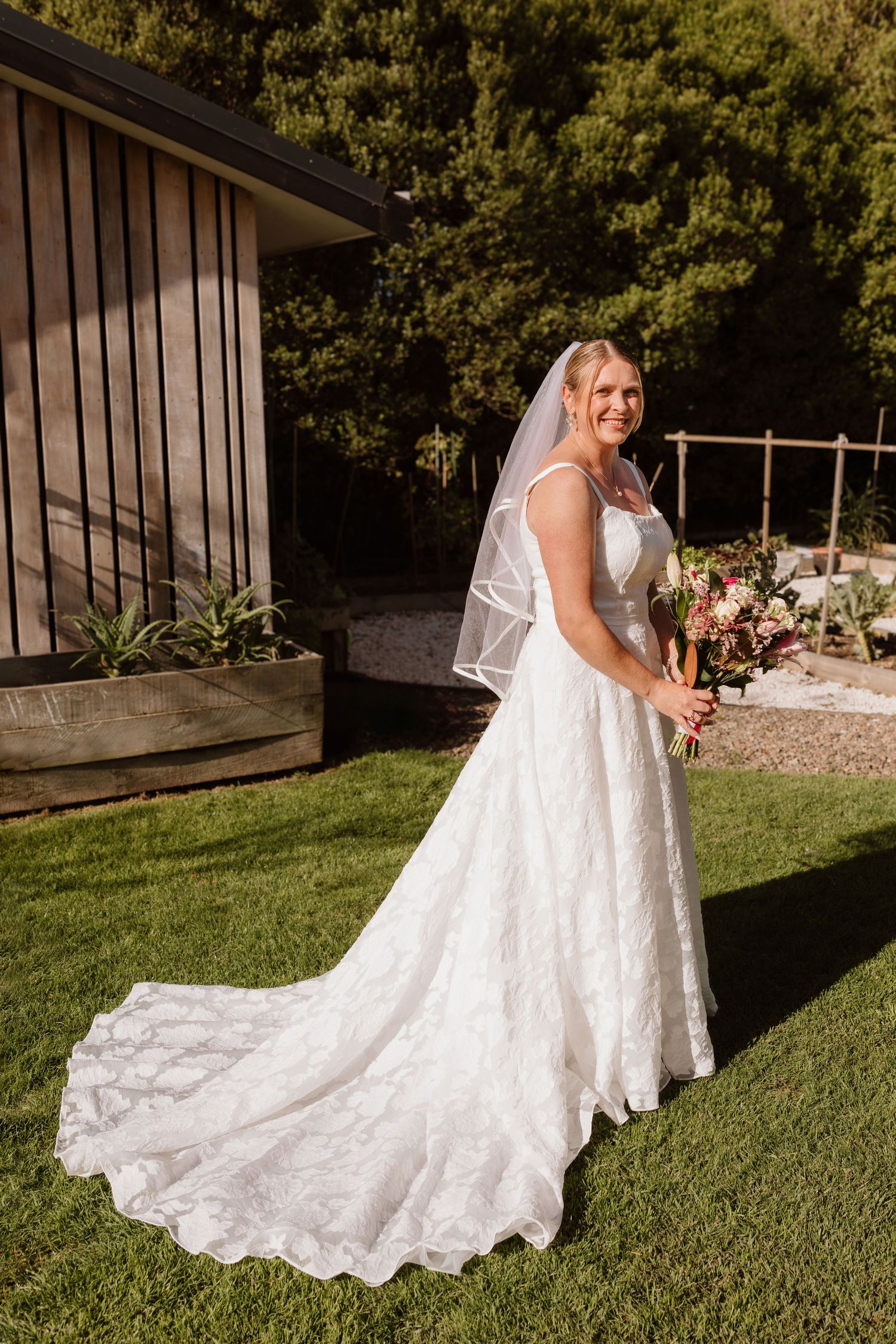 A bride in a flowing lace wedding gown with long train walks across a sunlit lawn on a beautiful day.