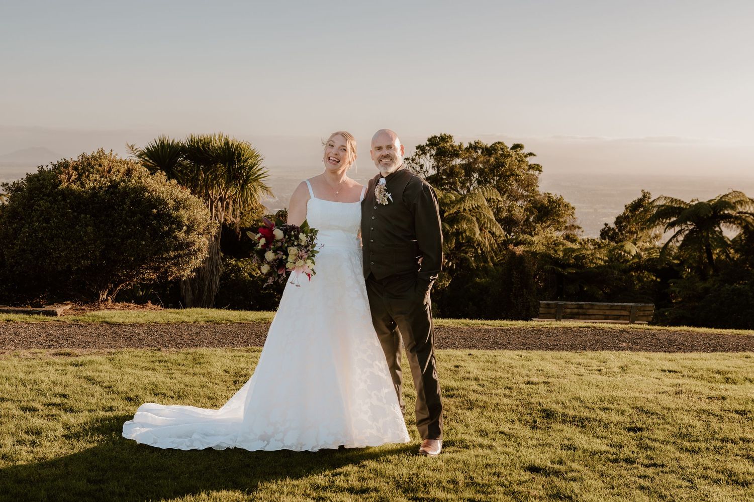 A couple in wedding attire poses on a hilltop at sunset with greenery and golden light in the background.