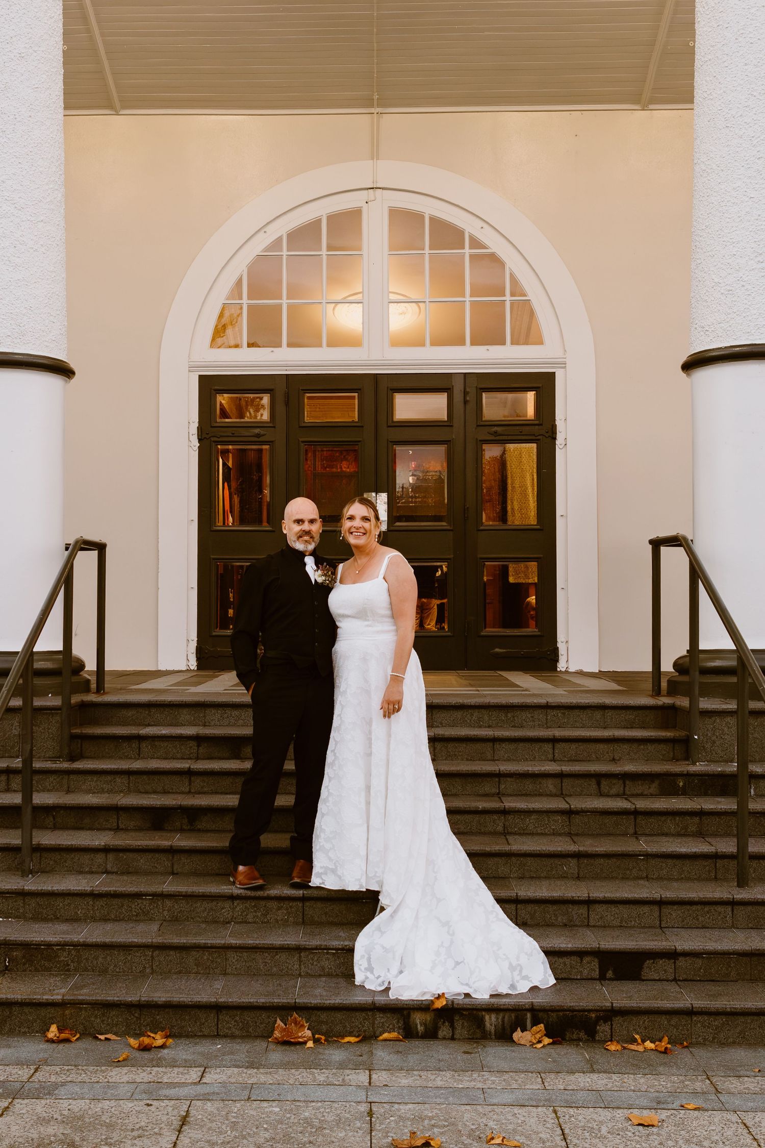 Wedding couple stands together on classic architecture steps with white columns in autumnal setting.