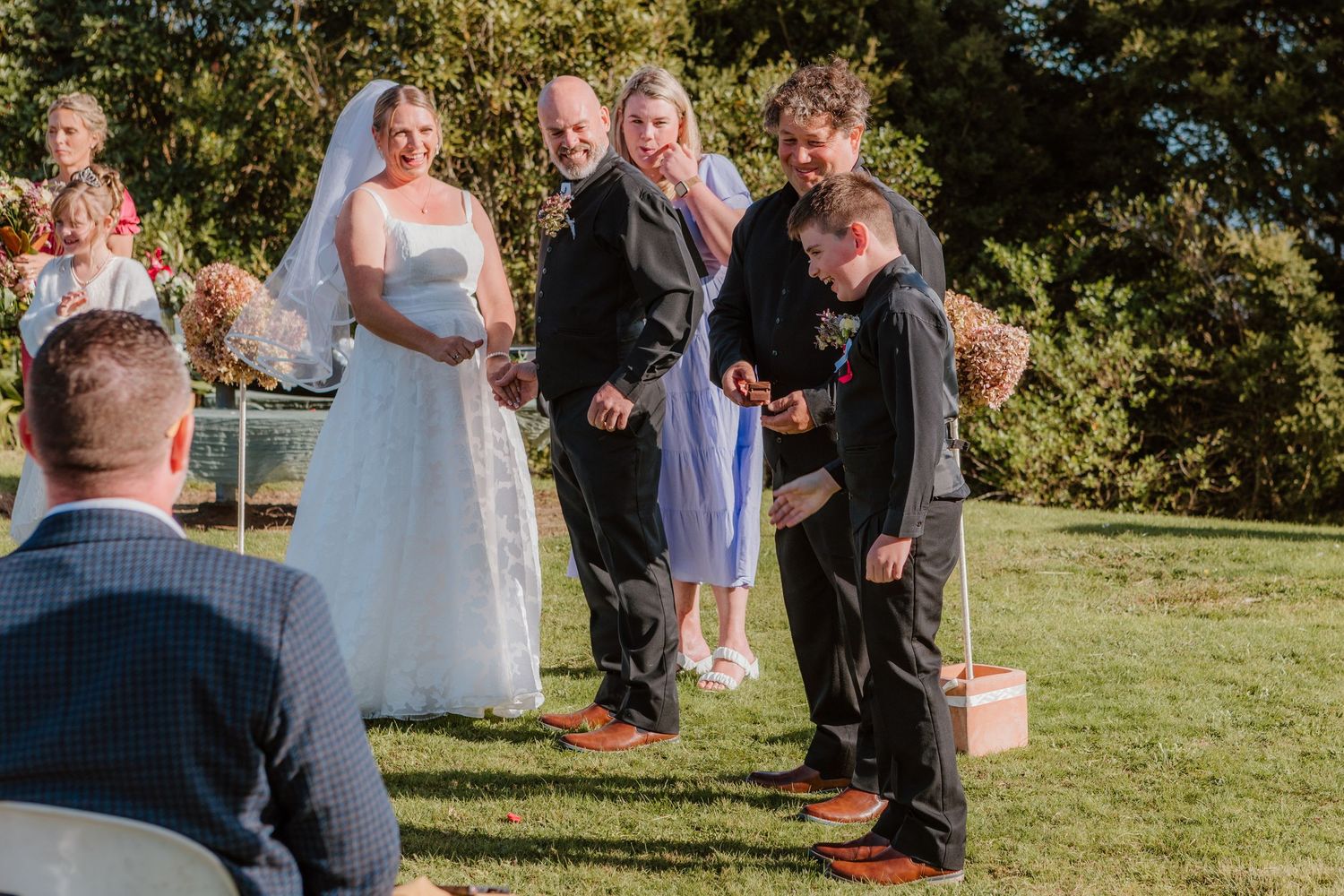An outdoor wedding ceremony takes place on a sunny day with guests gathered on a grassy lawn surrounded by trees.