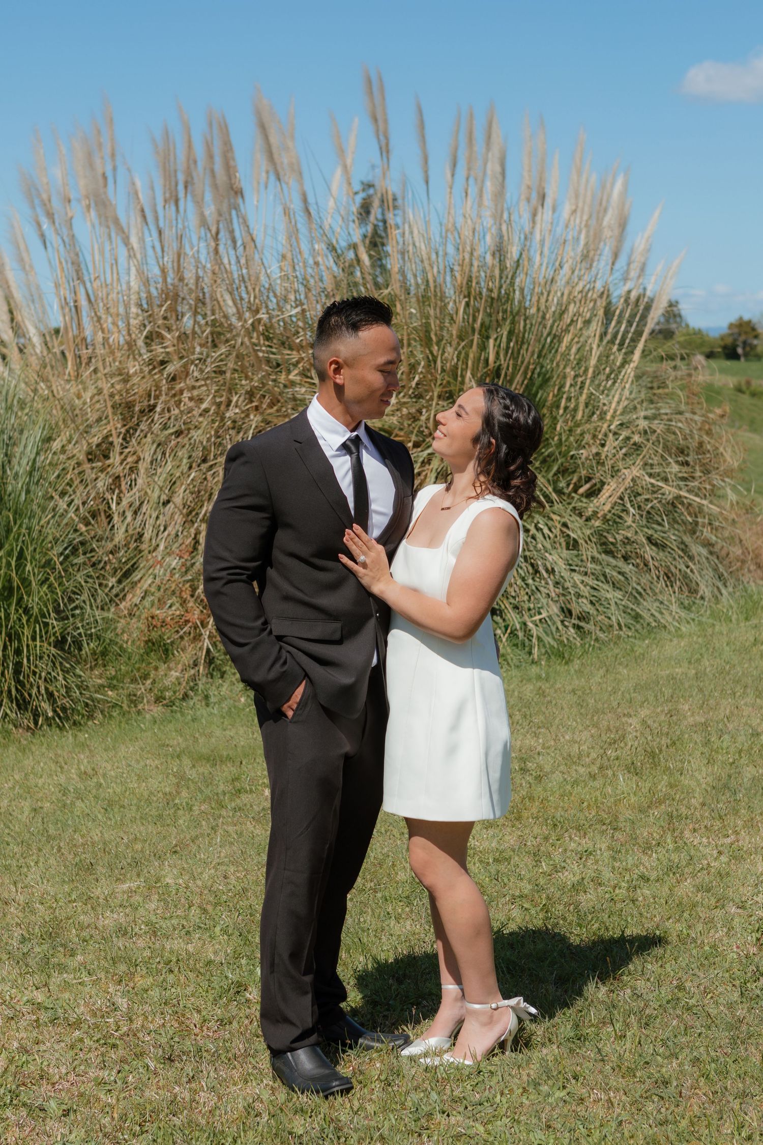 A couple in wedding attire share an intimate moment against tall ornamental grasses on a sunny day.