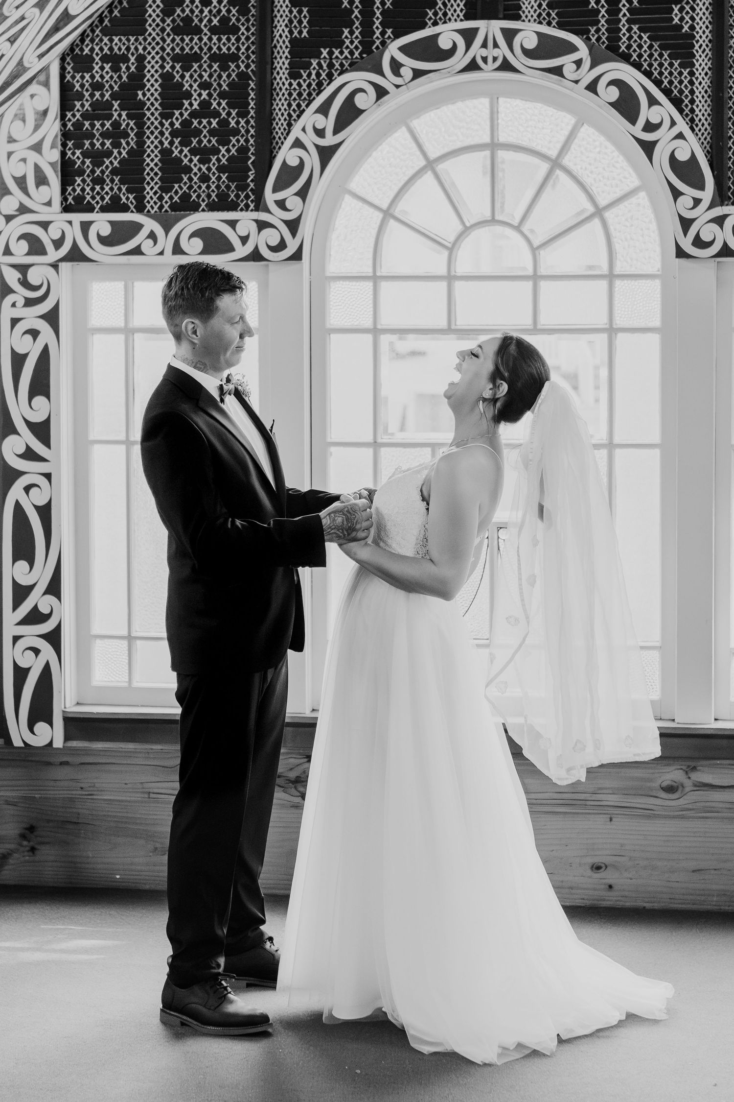 A romantic black and white wedding photo showing a couple in elegant attire standing by an ornate window.