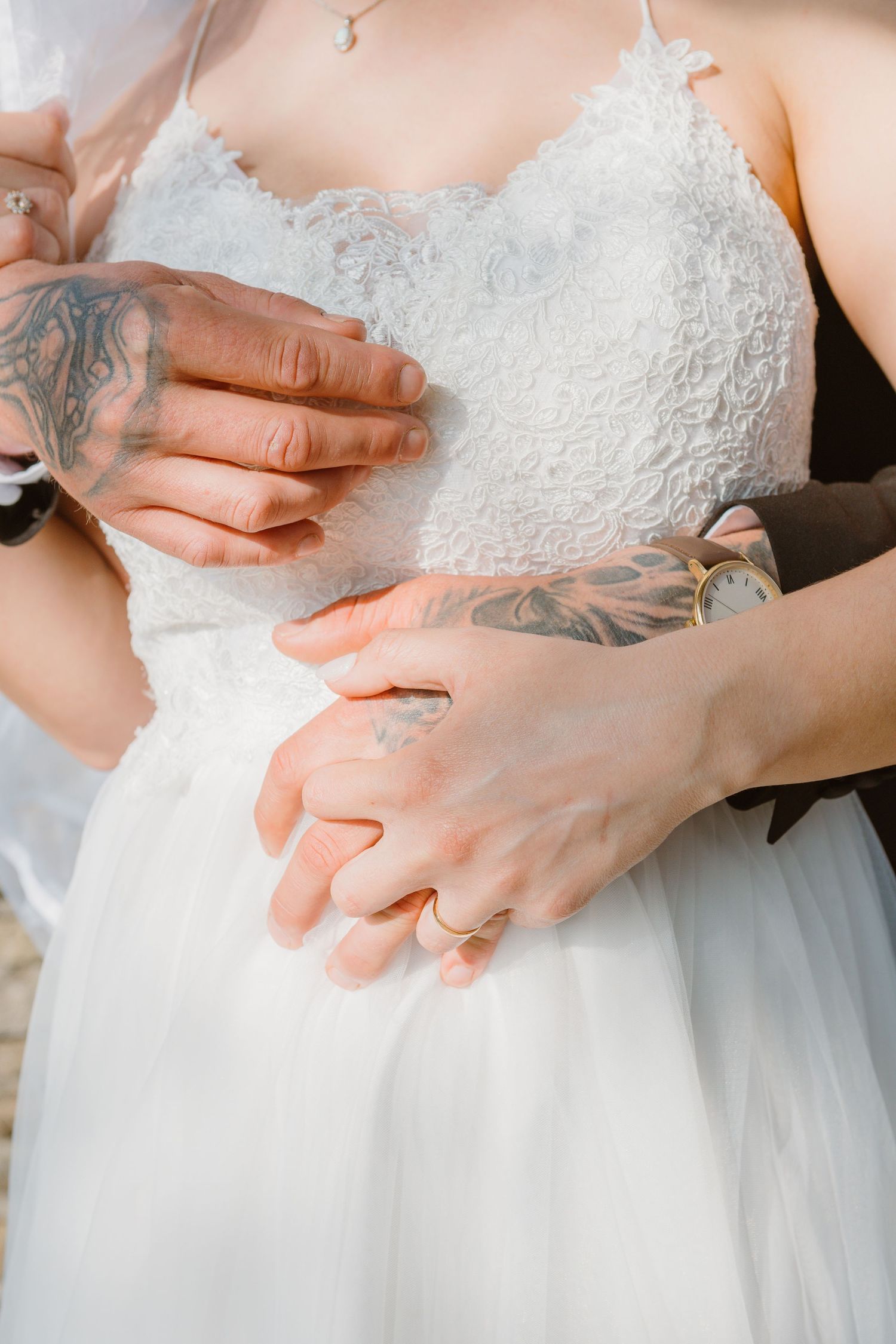 Closeup shots of hands with tattoos touching a white lace wedding dress bodice and tulle skirt.