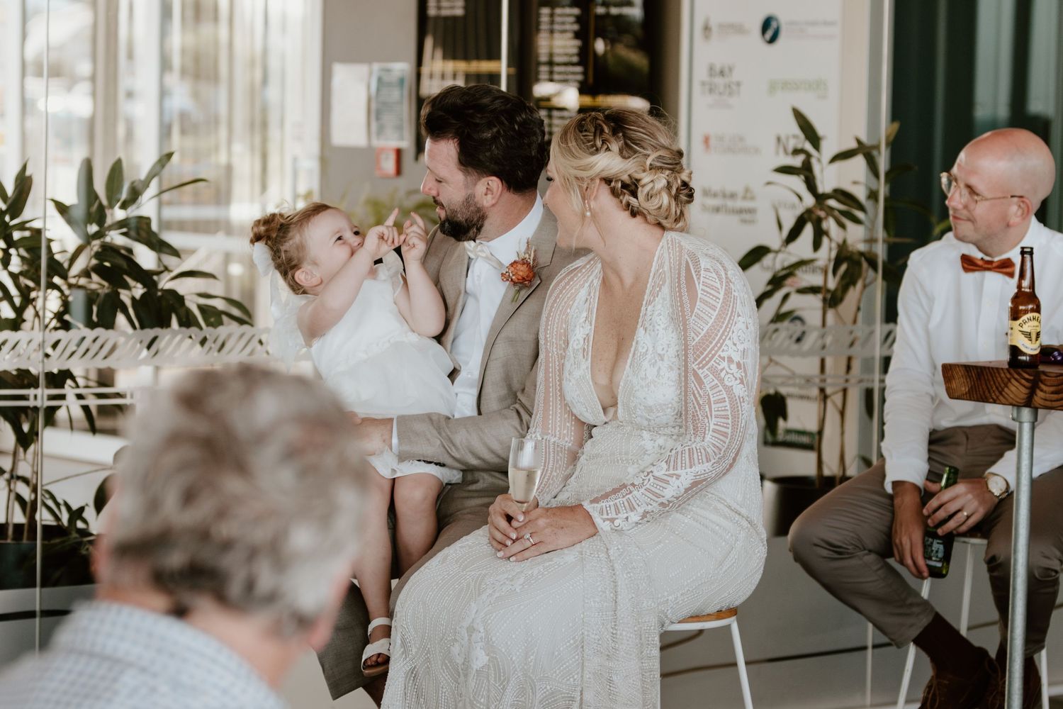 A bride in a lace gown sits at a cafe table while a small child is being held nearby.