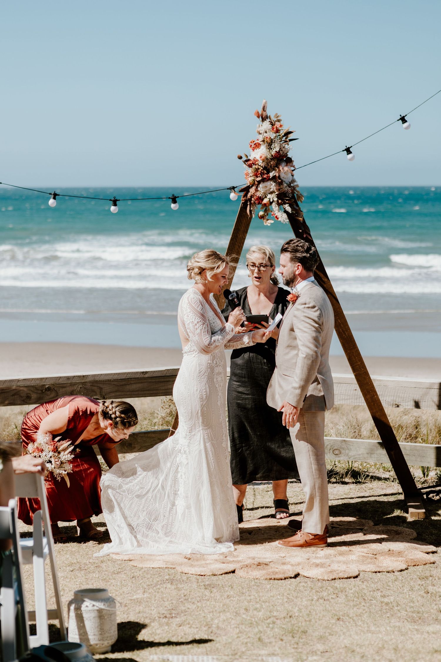 A beach wedding ceremony under a wooden arch decorated with pink flowers, with the ocean and string lights in the background.