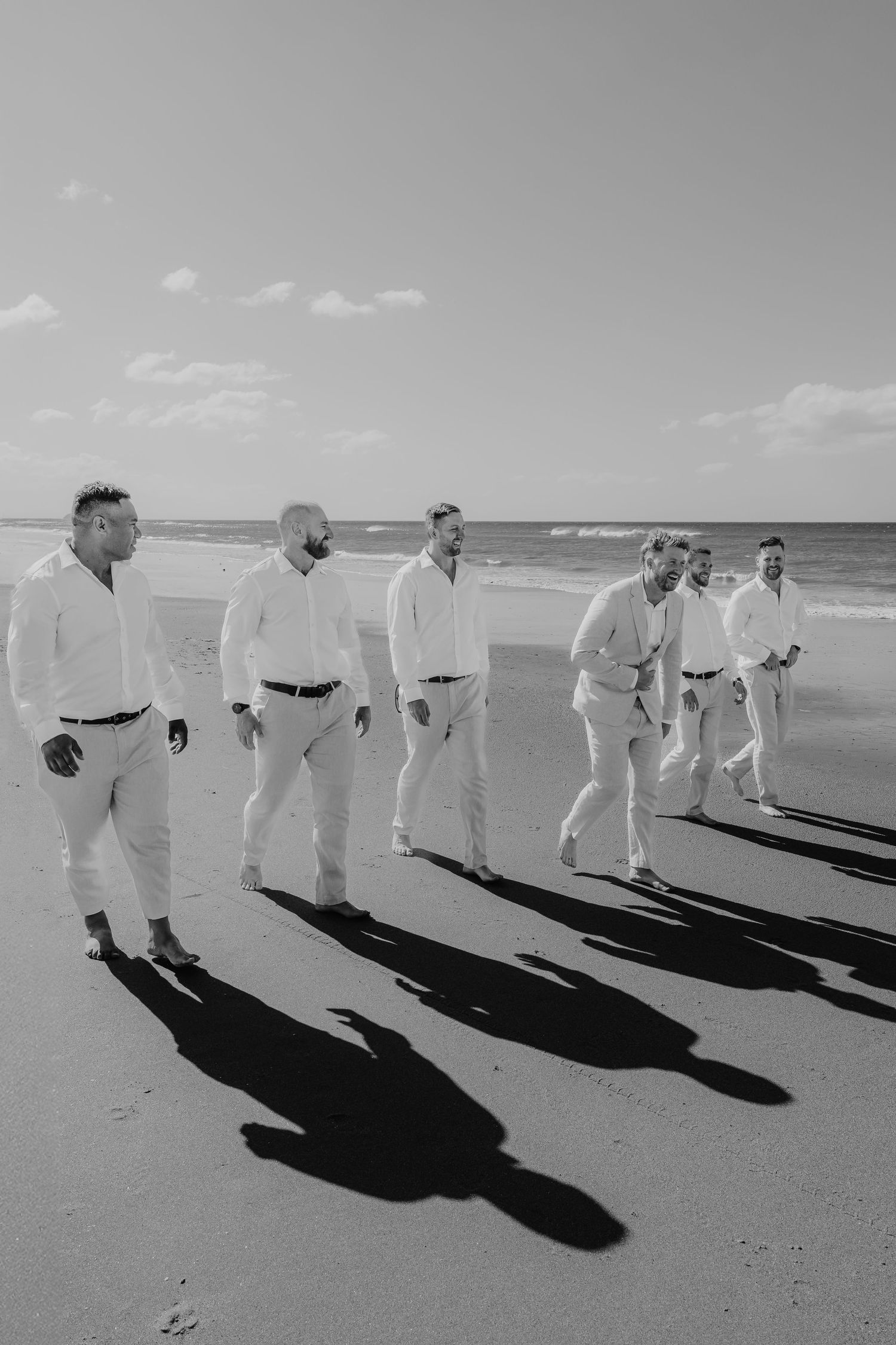 Black and white photo of groomsmen in white shirts walking on a beach, casting long shadows on the sand.