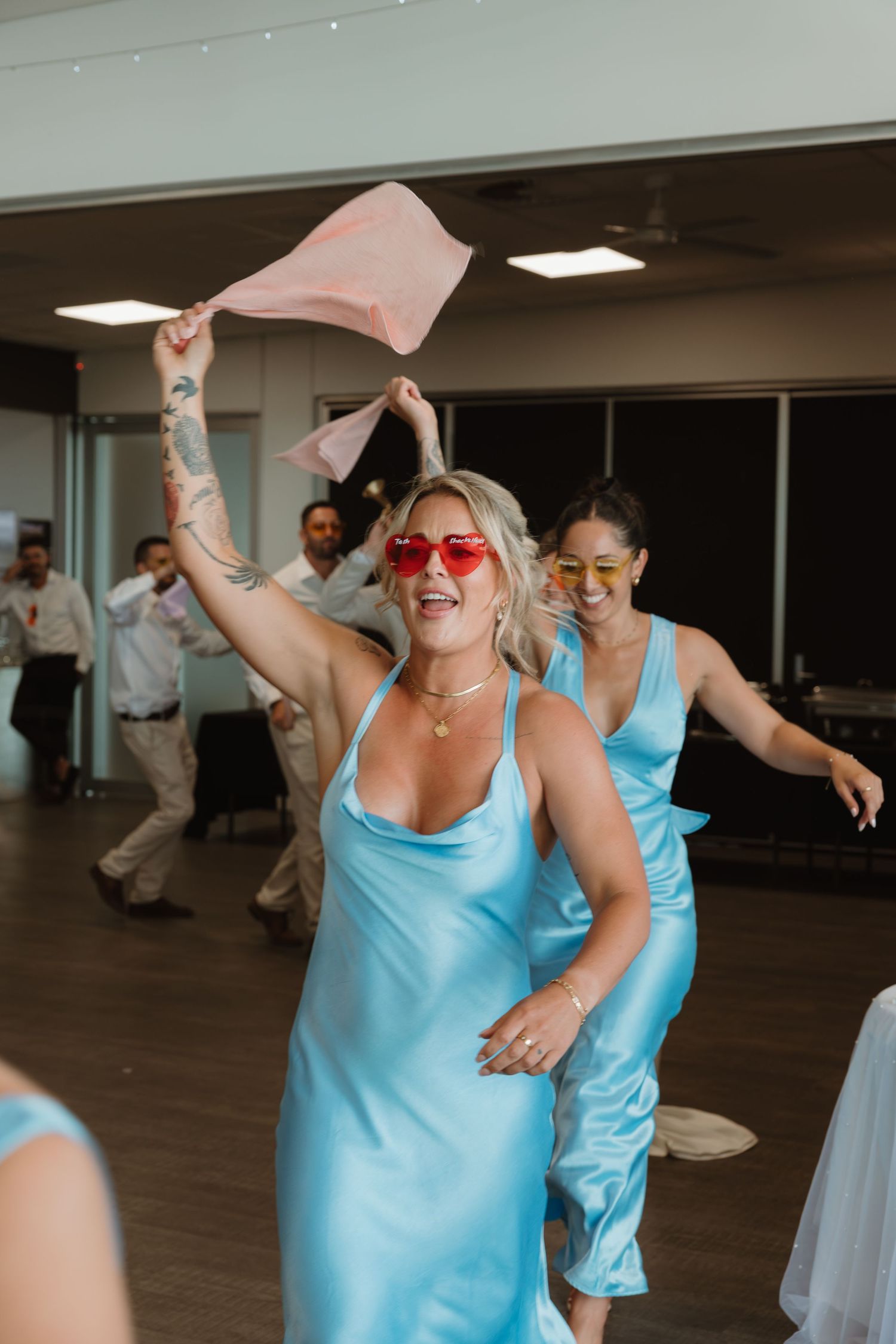 Bridesmaids in blue satin dresses dancing joyfully at a wedding reception, one wearing red heart sunglasses waving a pink cloth.