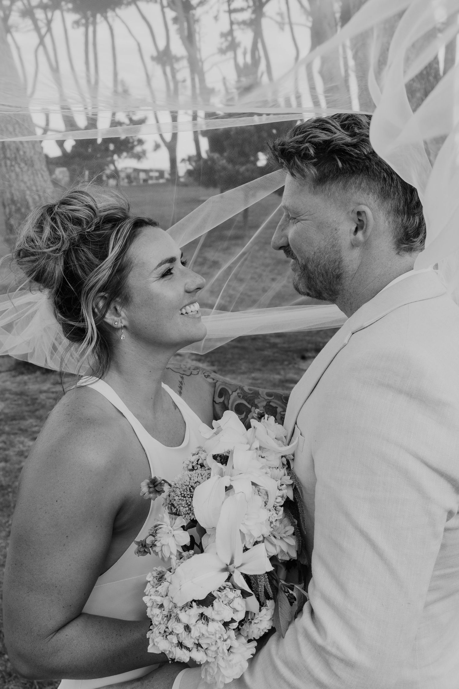 Black and white photo of a smiling bride holding a bouquet facing her groom outdoors at a wedding ceremony.