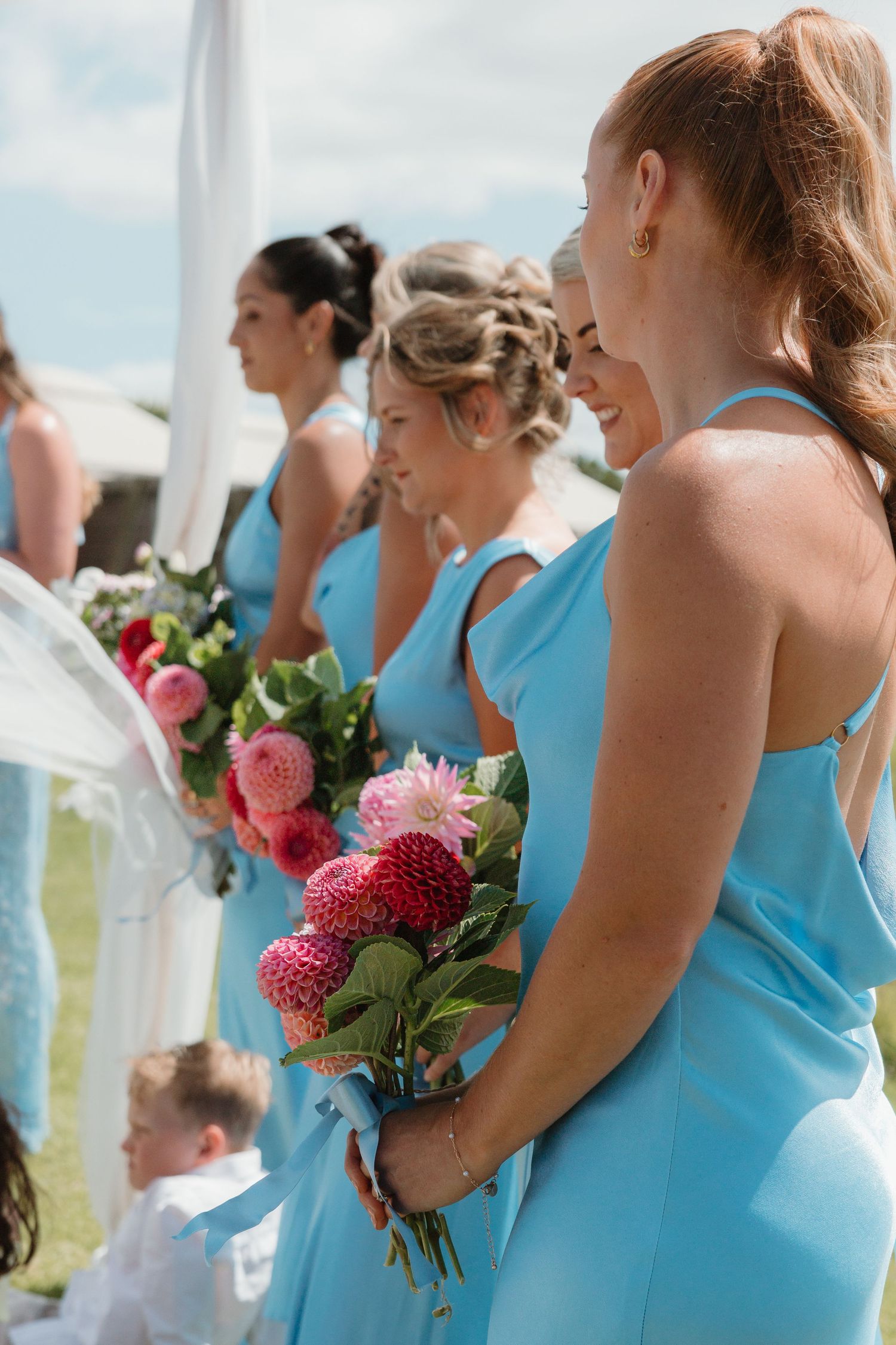 Bridesmaids in light blue dresses holding pink floral bouquets stand outdoors at a wedding ceremony.