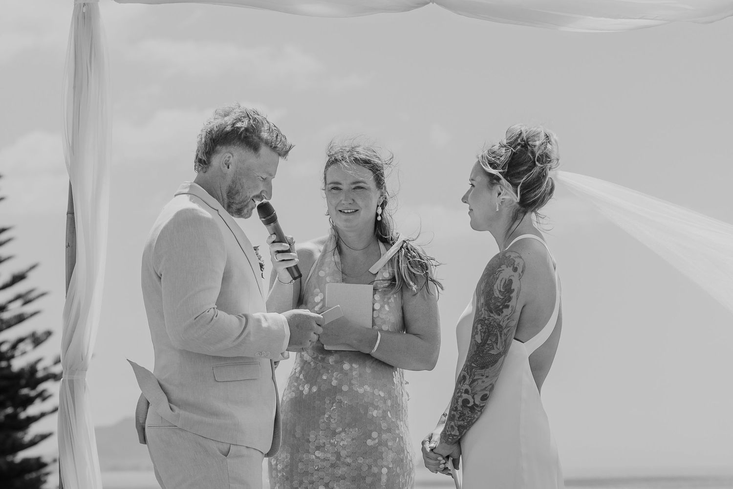 Black and white beach wedding ceremony with tattooed bride, groom in suit, and officiant smiling outdoors.