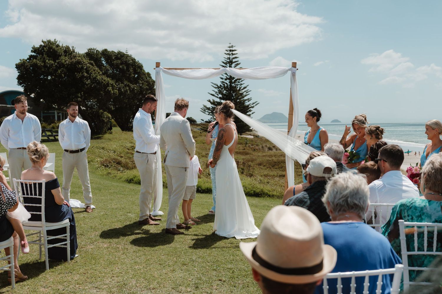 Outdoor beach wedding ceremony with couple exchanging vows under floral arch, guests seated on white chairs overlooking ocean.