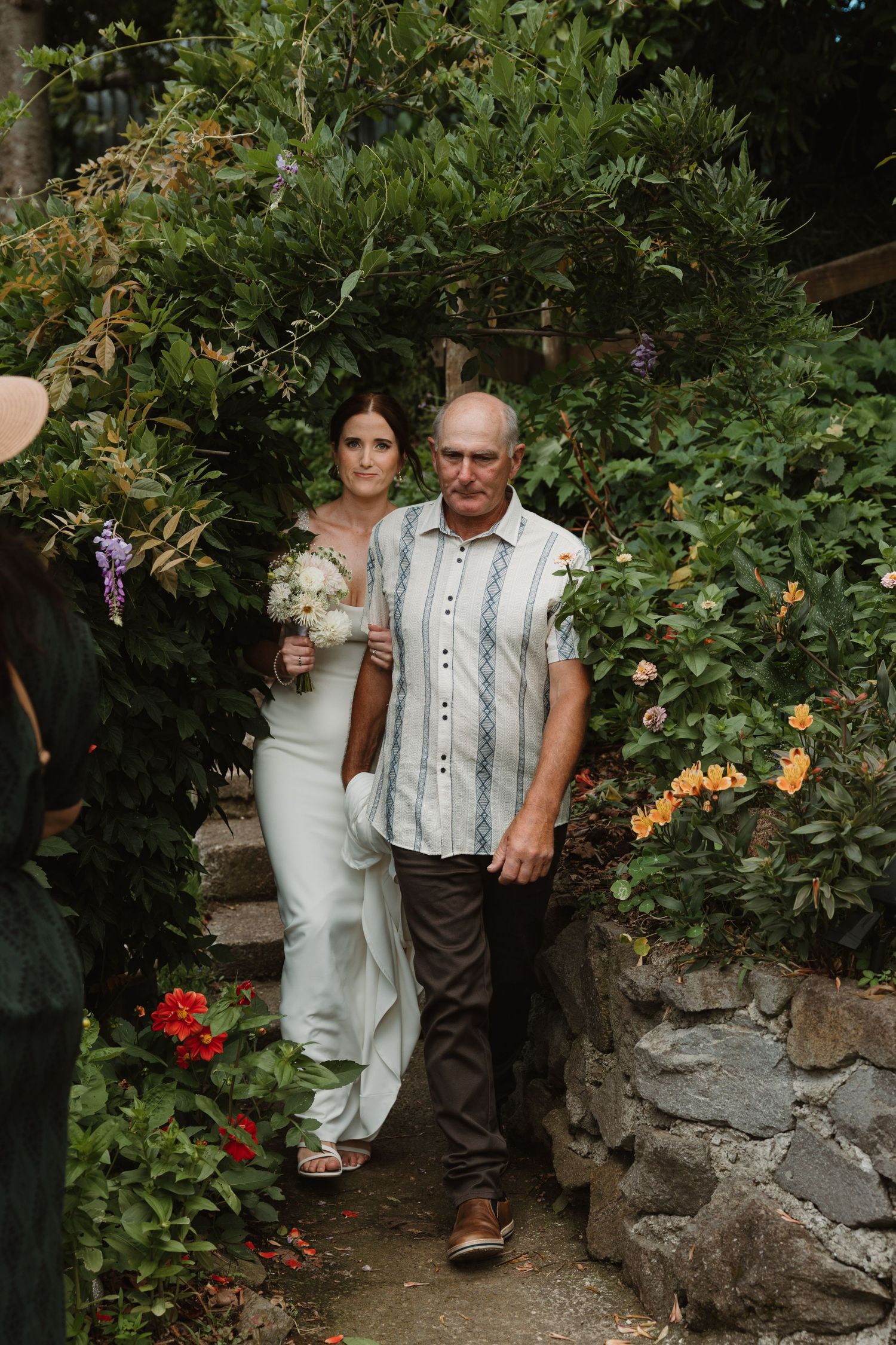 Bride in mint gown walks with older man through lush garden archway, holding white bouquet at outdoor wedding ceremony.