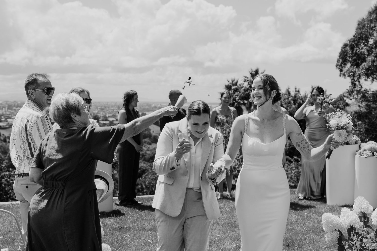 Black and white photo of two brides celebrating after their wedding ceremony outdoors with guests cheering around them.