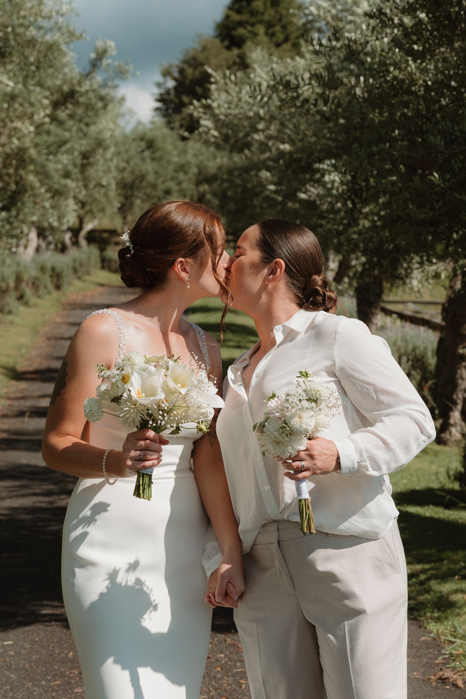 Two brides in white attire kiss while holding bouquets on a tree-lined path on their wedding day.