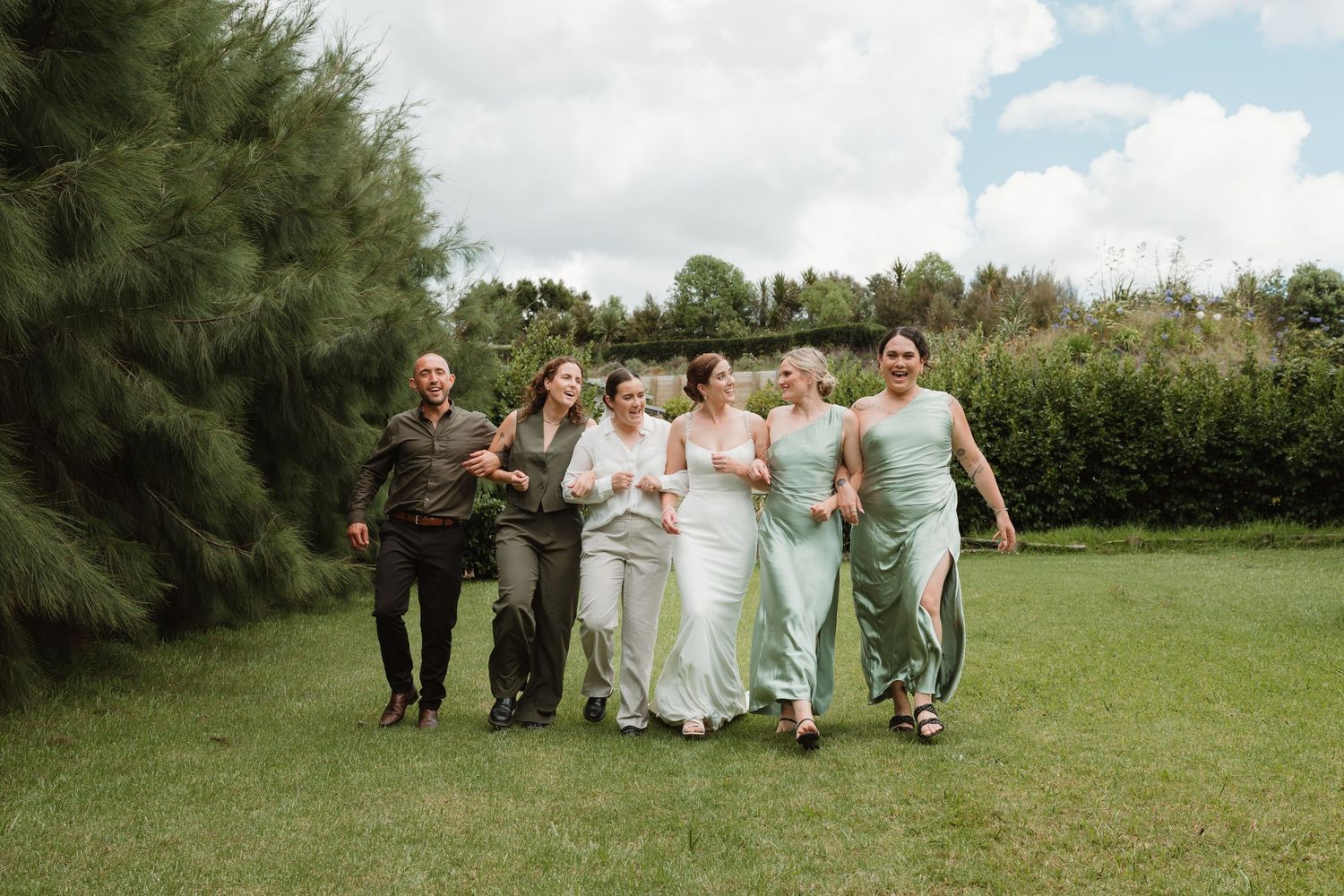 A joyful wedding party walks across a green lawn, with the bride in white and bridesmaids in sage green dresses.