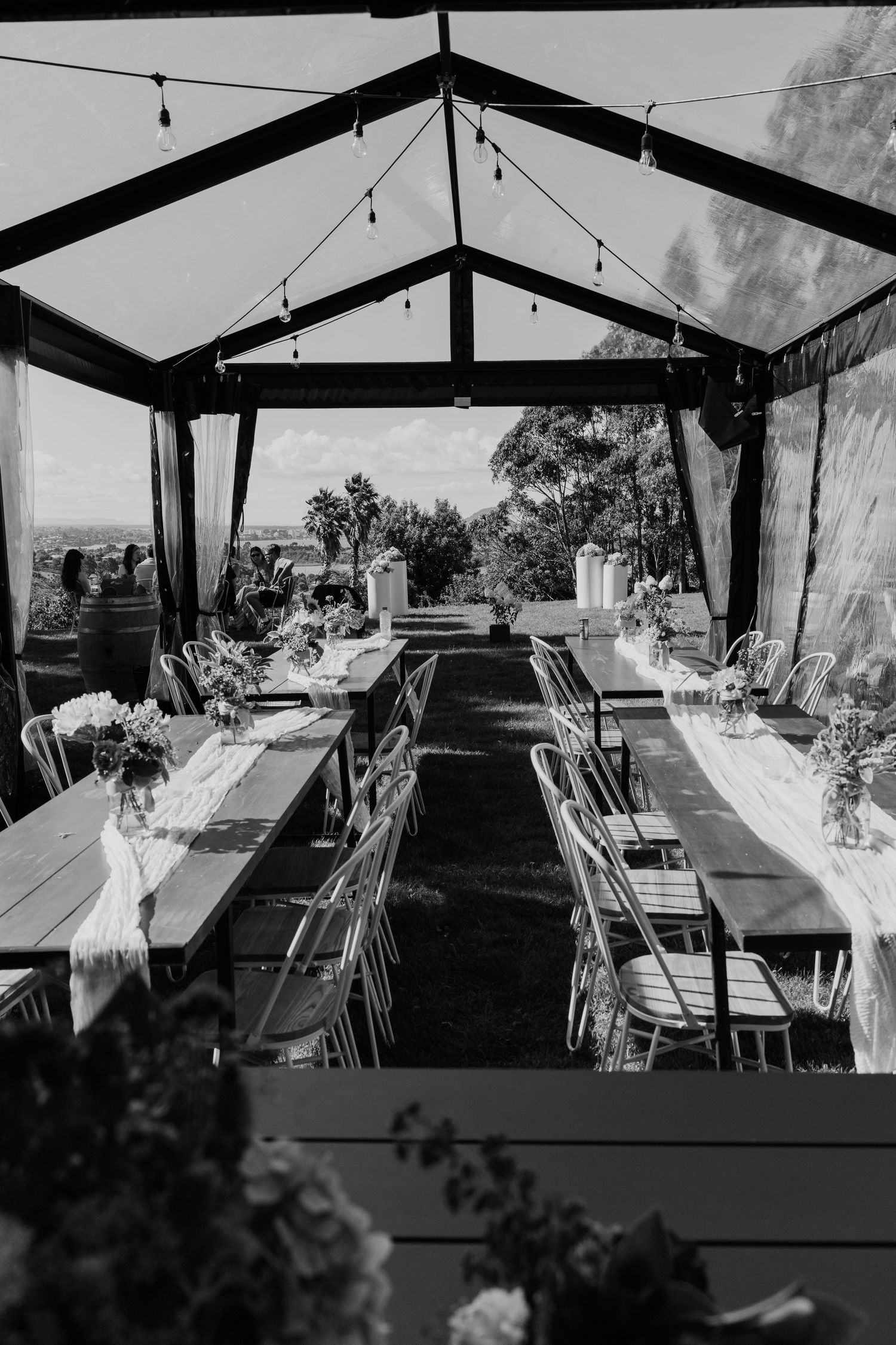 Black and white photo of an elegant outdoor wedding reception venue with long tables, floral arrangements, and string lights.