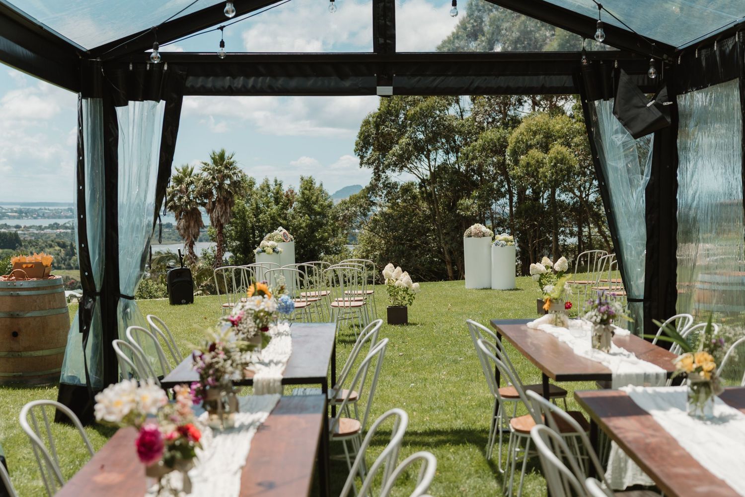 Outdoor wedding reception under a glass pavilion with long wooden tables, floral centerpieces, and a lush green garden backdrop.
