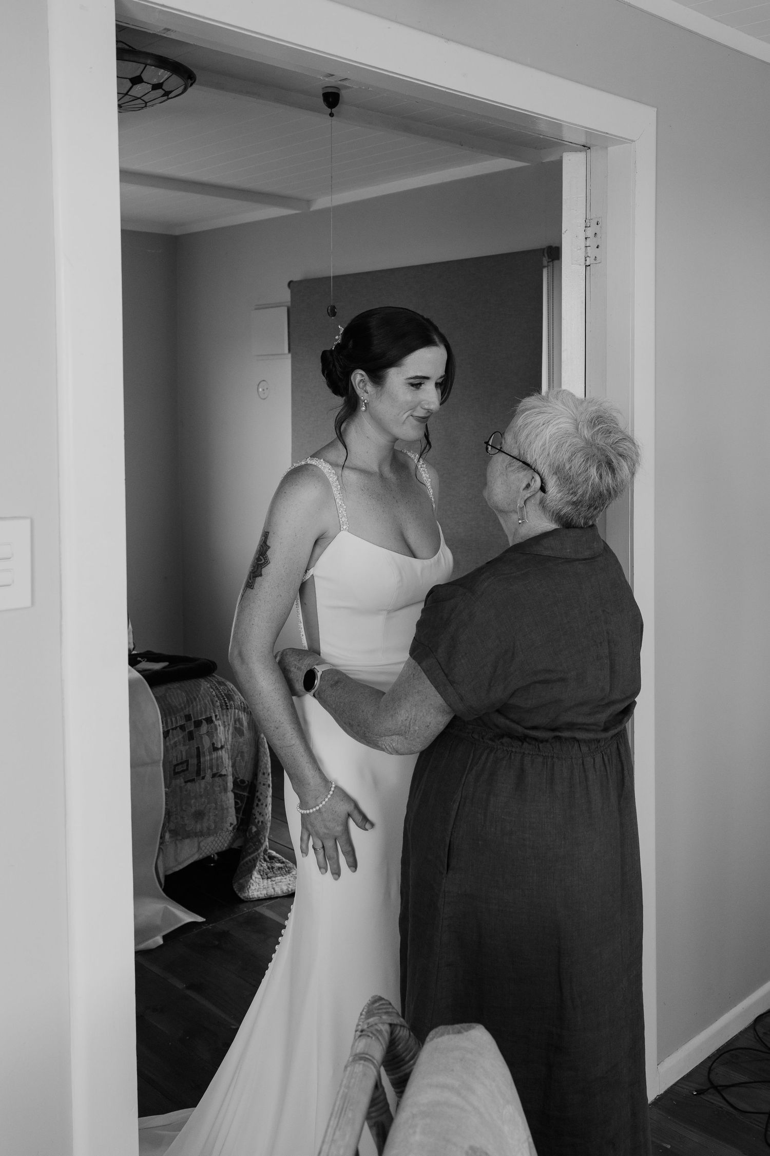 Black and white photo of a bride in a white gown being helped by an elderly woman in a doorway.