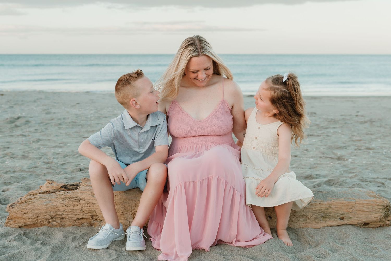 Pregnant woman in pink dress sits on driftwood with two children on a beach, sharing a tender family moment.