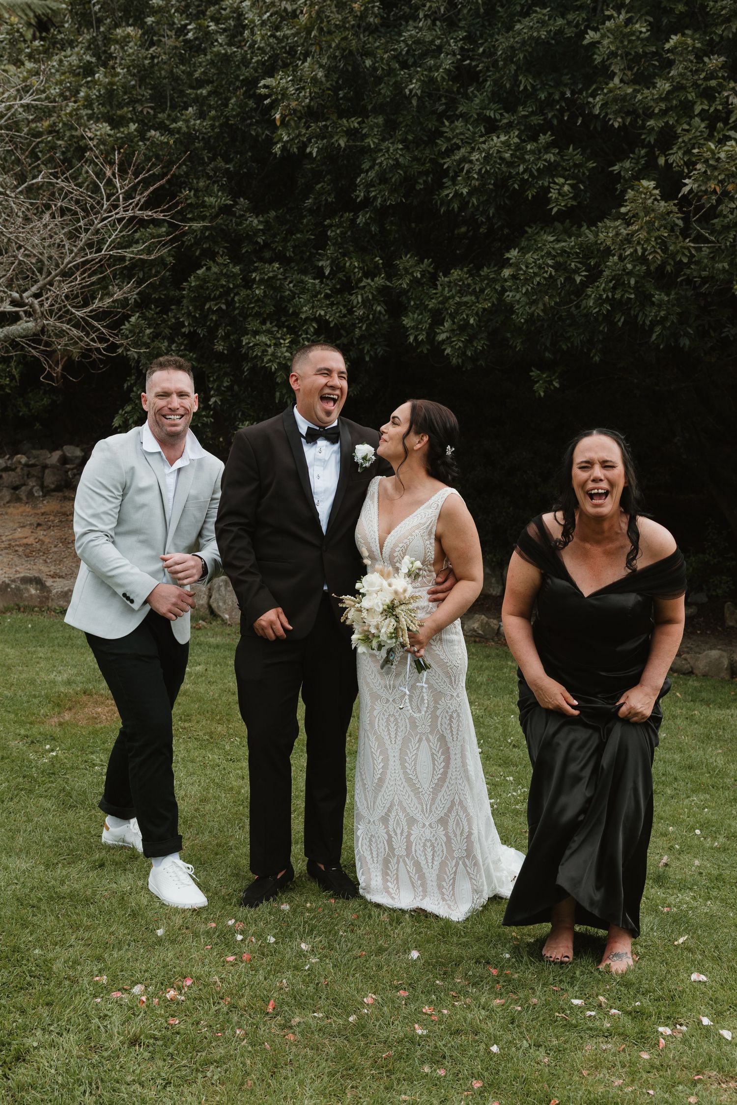 Wedding party members share a candid laughing moment together on a grassy lawn during outdoor photo session.