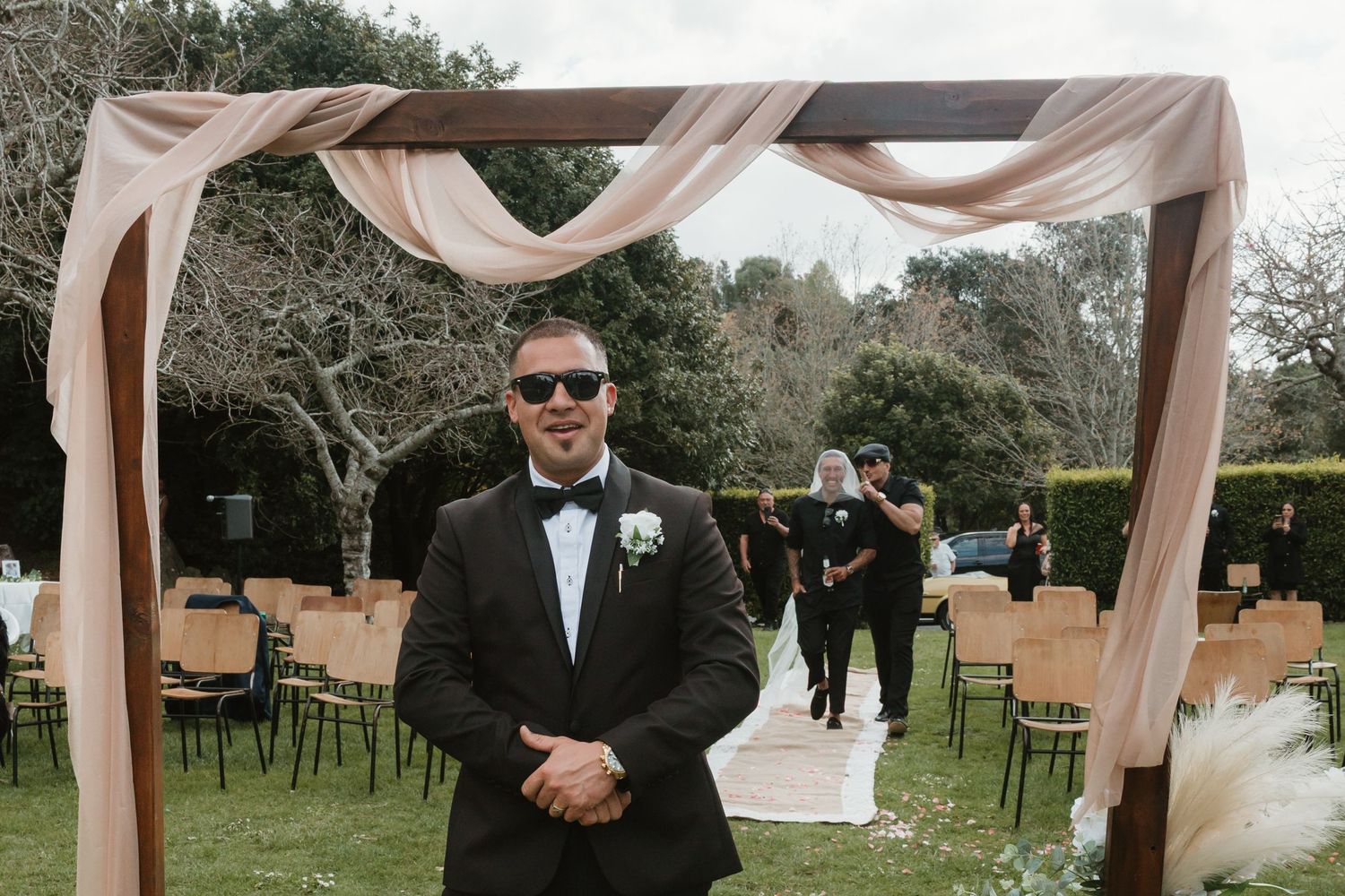 Wedding ceremony guests gather outdoors under elegant blush-colored fabric draping between trees at a garden venue.