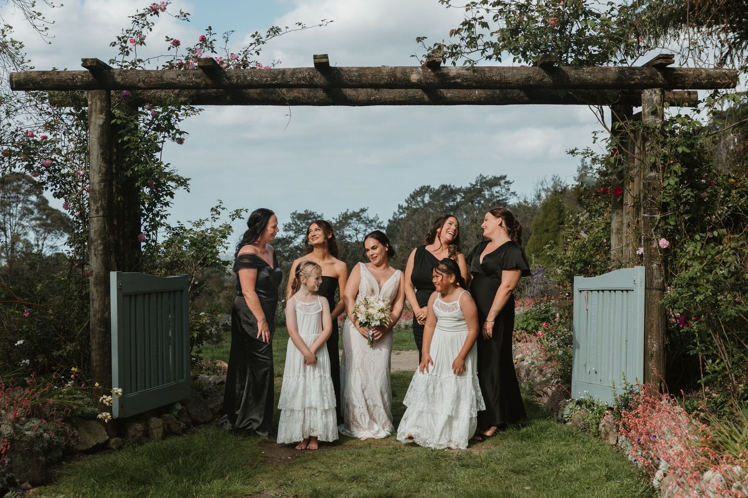 Wedding party poses outdoors in front of rustic wooden fence and lush greenery during summer celebration.