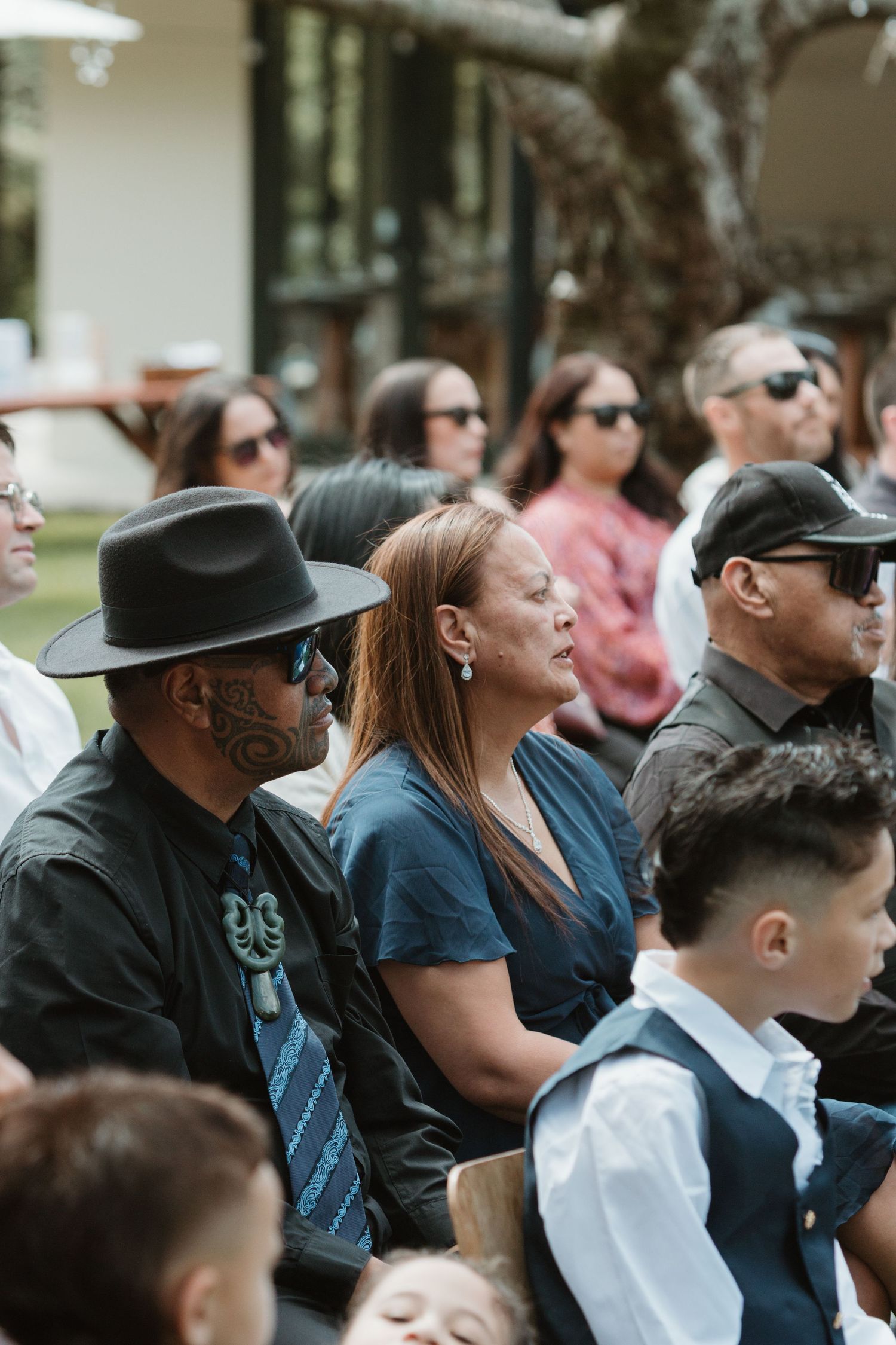 A crowd of spectators wearing sunglasses sits outdoors watching an event with engaged expressions.